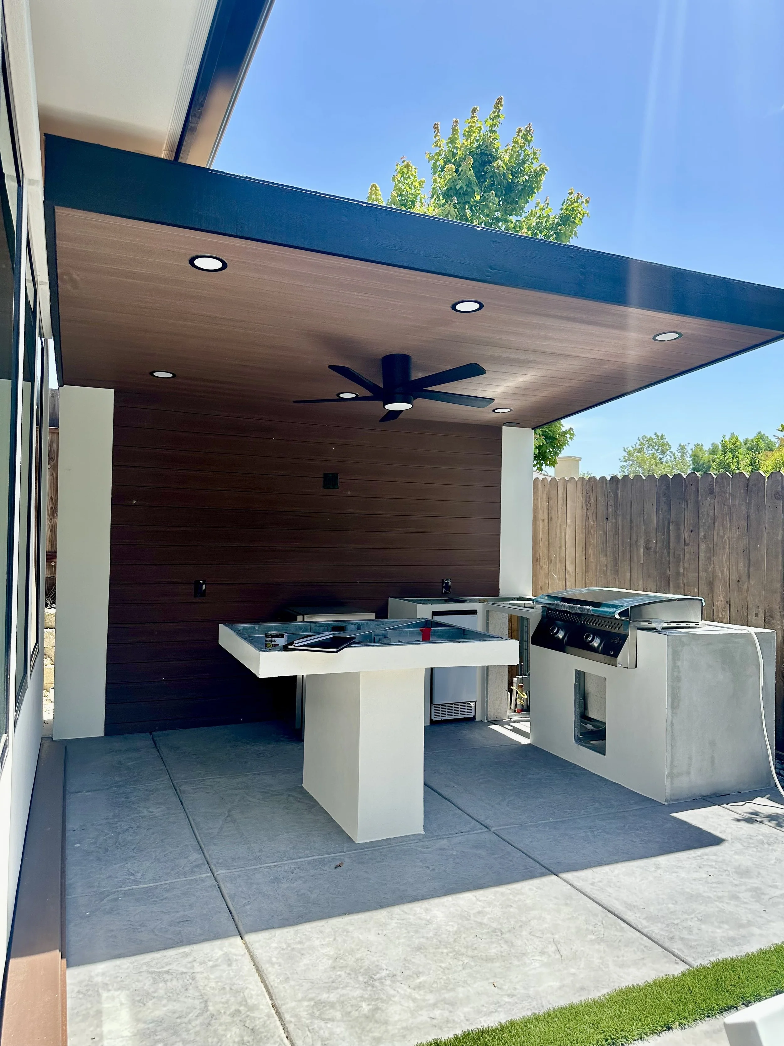 Outdoor patio area with built-in grill, ceiling fan, and a wooden ceiling. There is a counter with a stovetop, a small refrigerator, and a wooden privacy fence. Green trees and a blue sky are visible in the background.