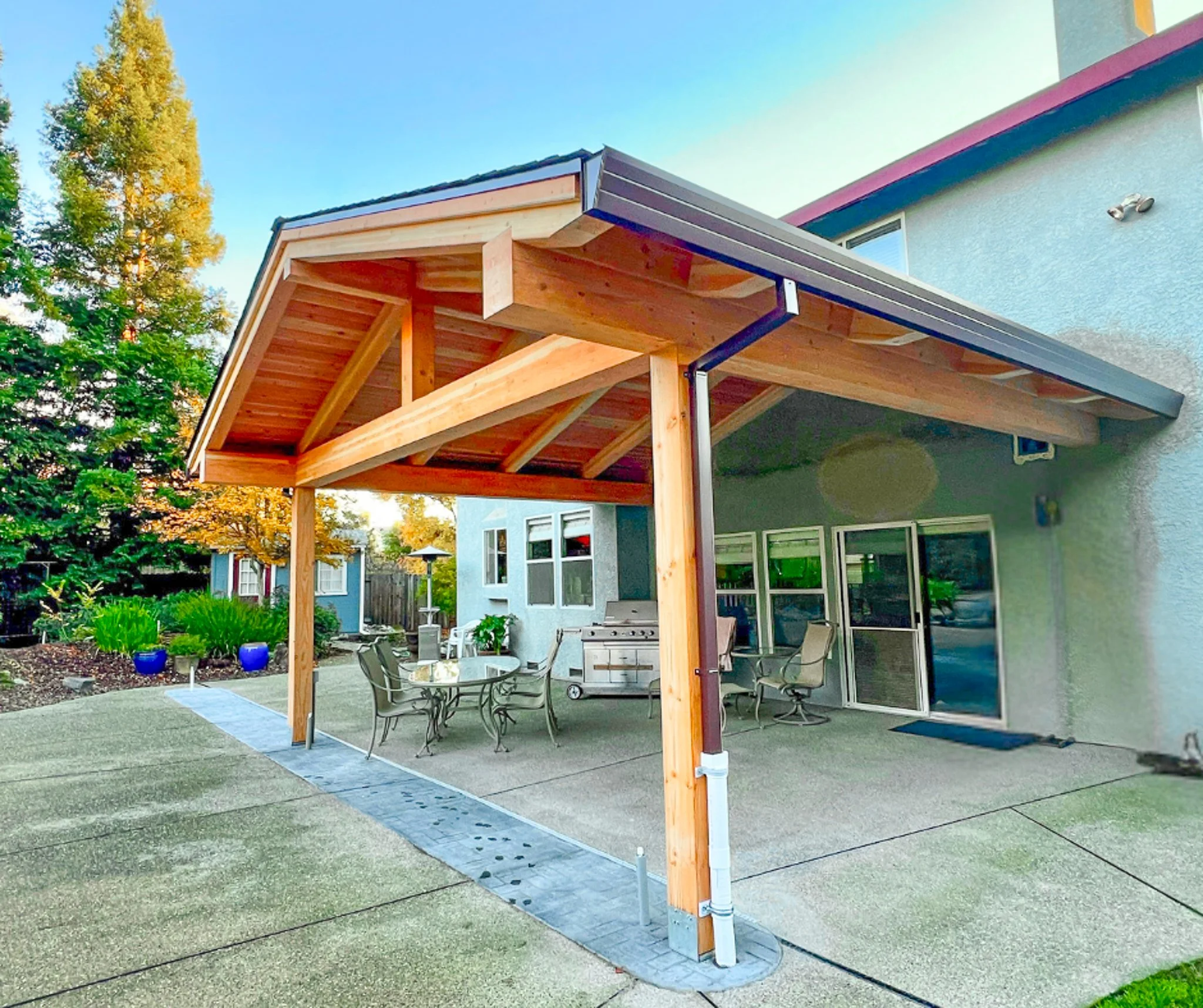 Backyard patio with new wooden pergola, patio table, chairs, barbecue grill, and potted plants, overlooking neighboring houses and trees.
