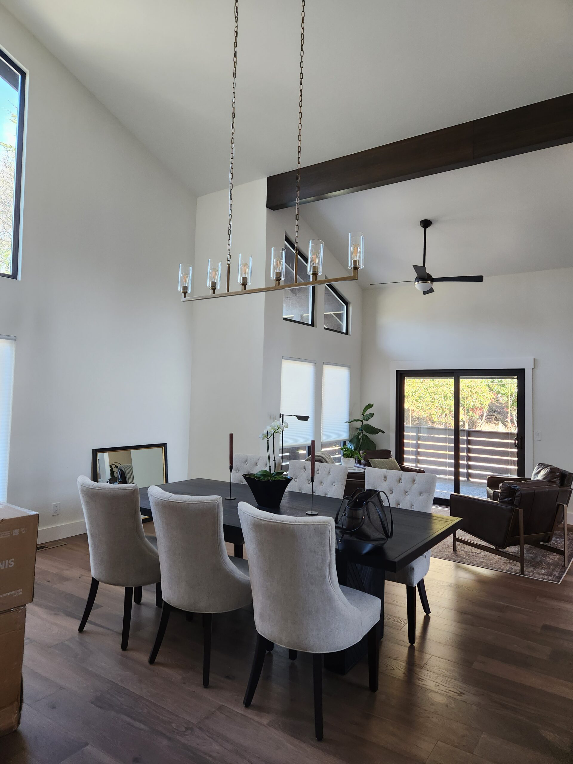 Modern dining and living room with high ceilings, white walls, large windows, and wooden flooring, featuring a dark dining table with beige chairs, a black chandelier, and a sitting area with armchairs and a sliding glass door.