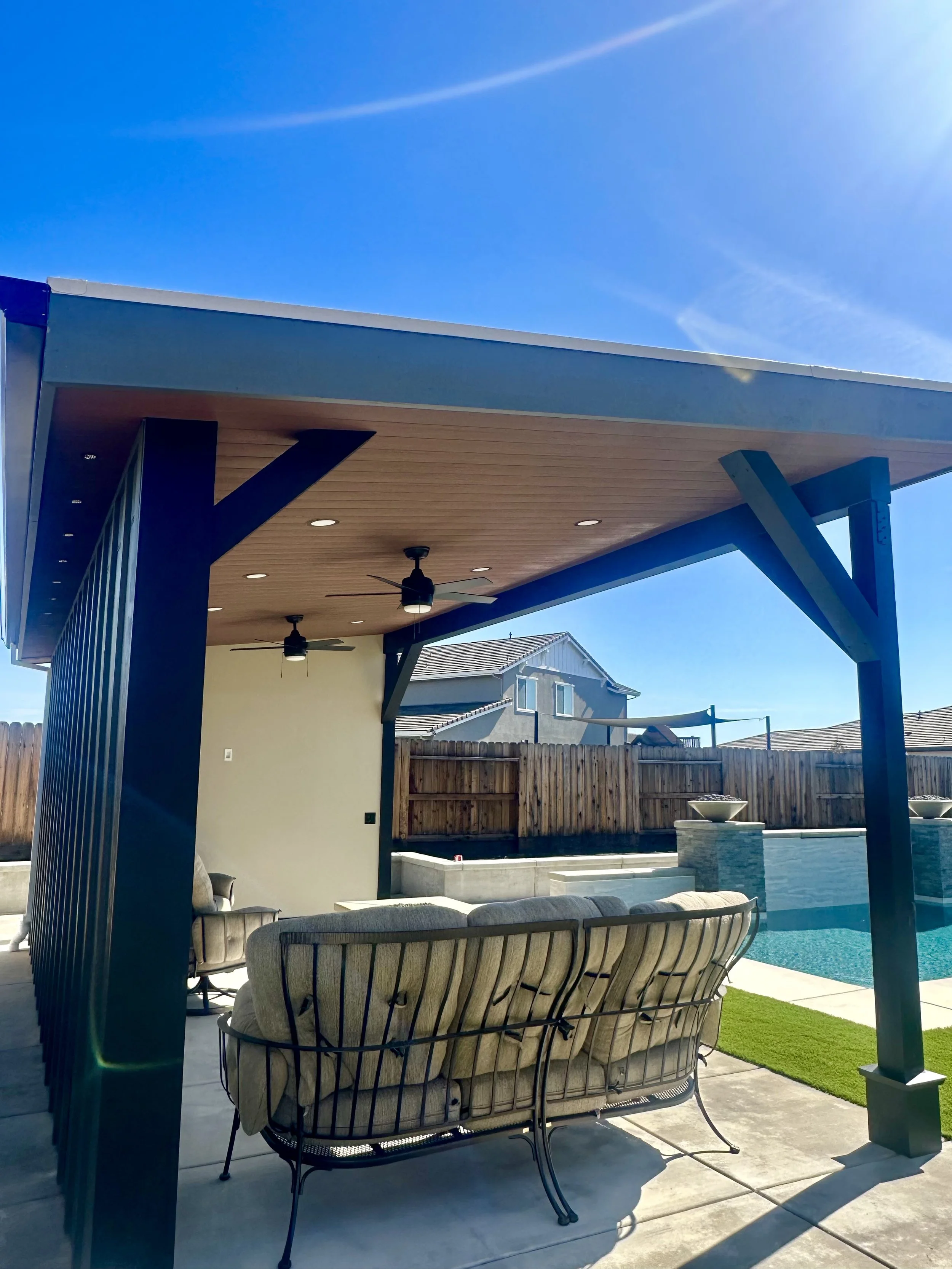 Backyard patio area with a covered seating space, ceiling fans, and a view of a swimming pool and a neighboring house, under a clear blue sky.