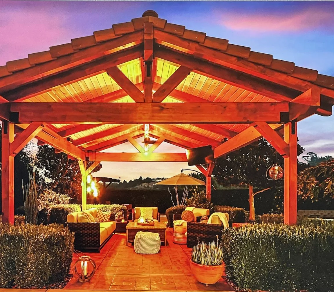 An outdoor wooden pavilion with string lights illuminating a seating area with yellow couches, armchairs, and a coffee table, set against a sunset sky.