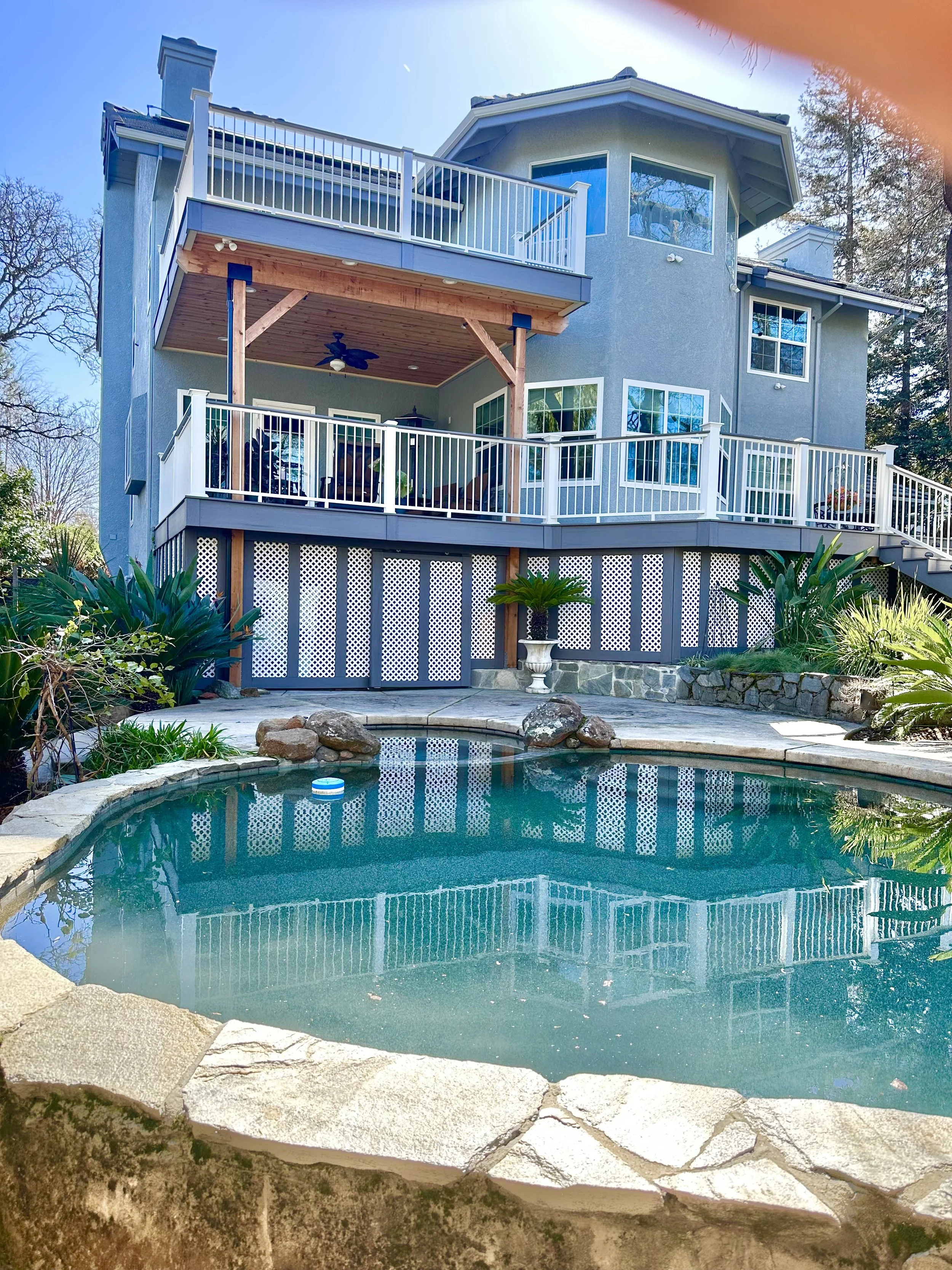 A multi-story house with gray exterior walls and large windows, featuring balconies with white railings and a wooden porch. In front, there's a backyard with a small stone-edged swimming pool surrounded by rocks and plants.