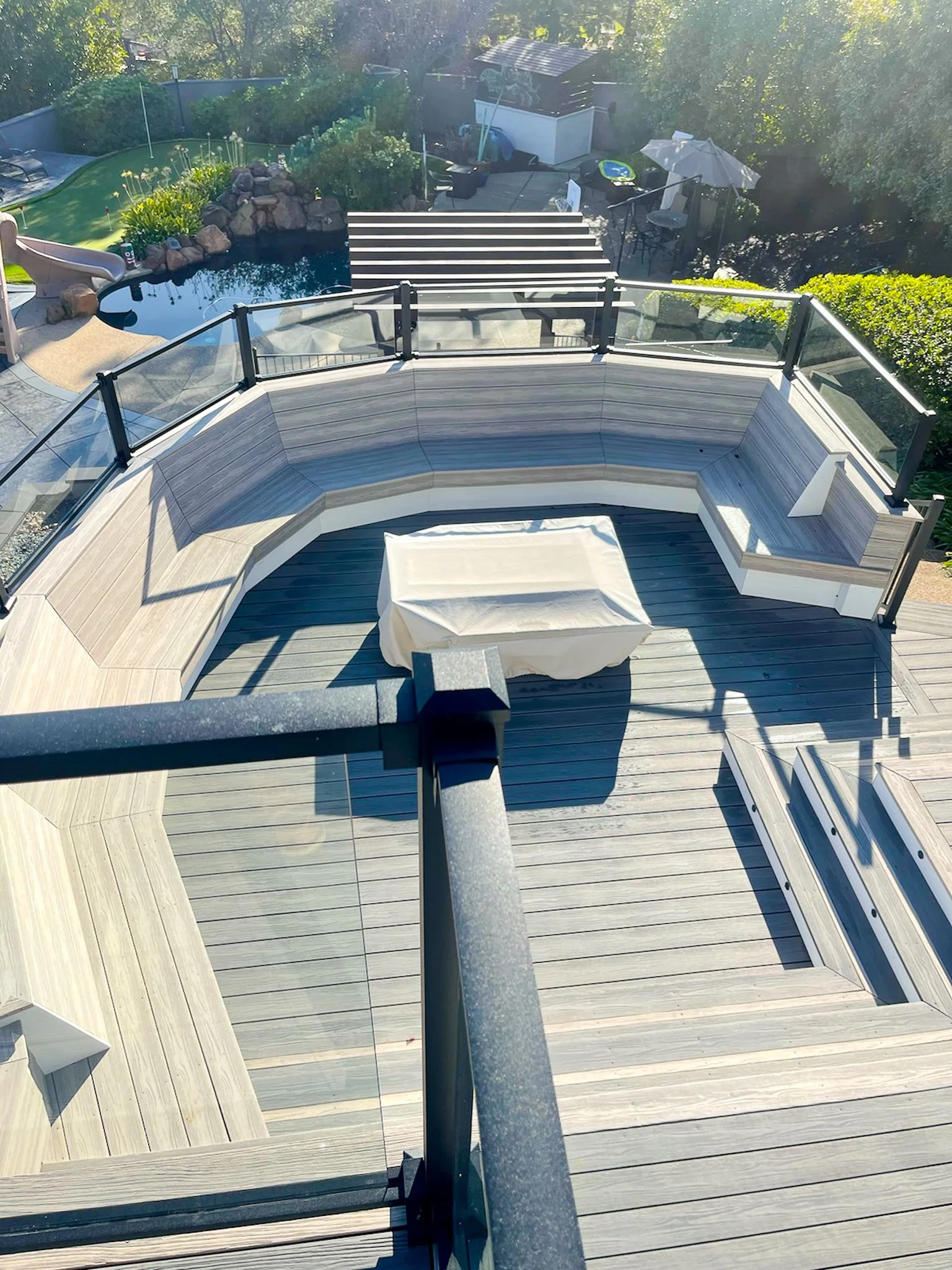View of an outdoor balcony with a covered table, surrounded by glass railings. The deck has wood planks and stairs leading down to a backyard with a small pool, artificial grass, patio furniture, and gardening elements.