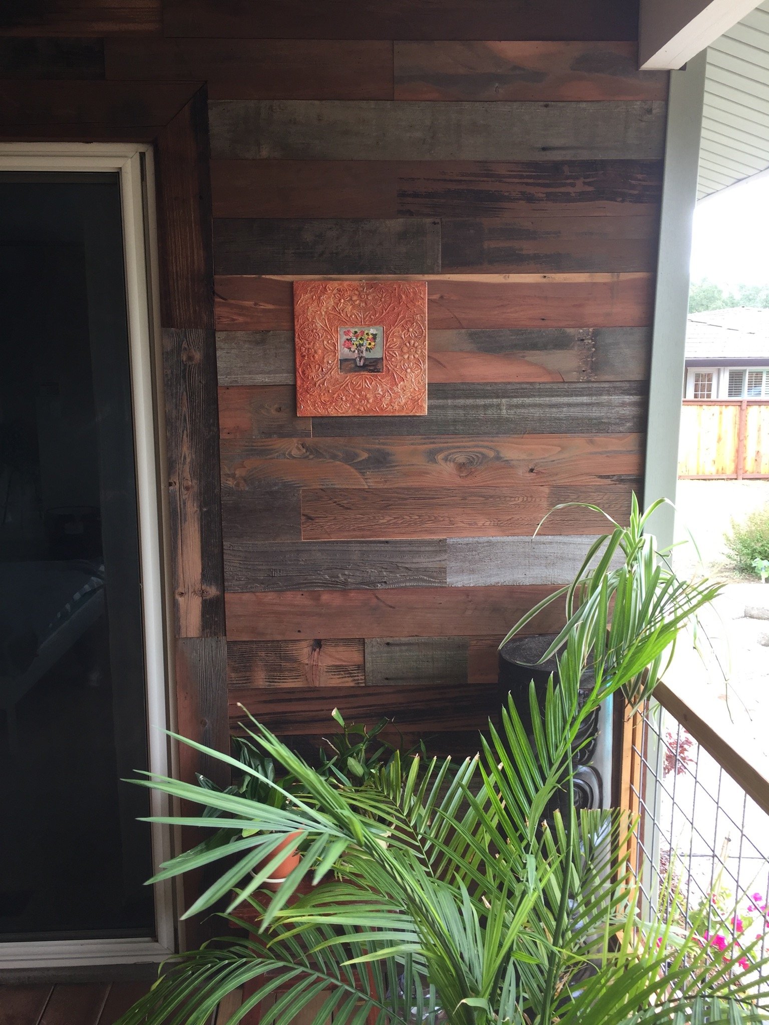 A balcony corner with a wooden wall, a small colorful painting of flowers in a vase, potted plants, and a sliding glass door.