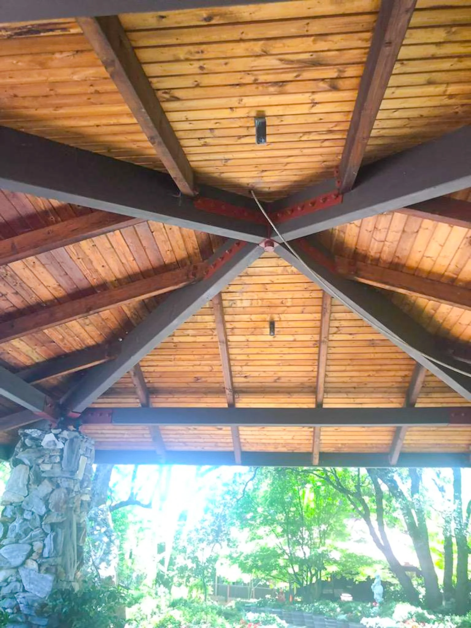 The ceiling of a gazebo or pavilion with wooden planks and supporting beams, outdoor garden visible in the background.
