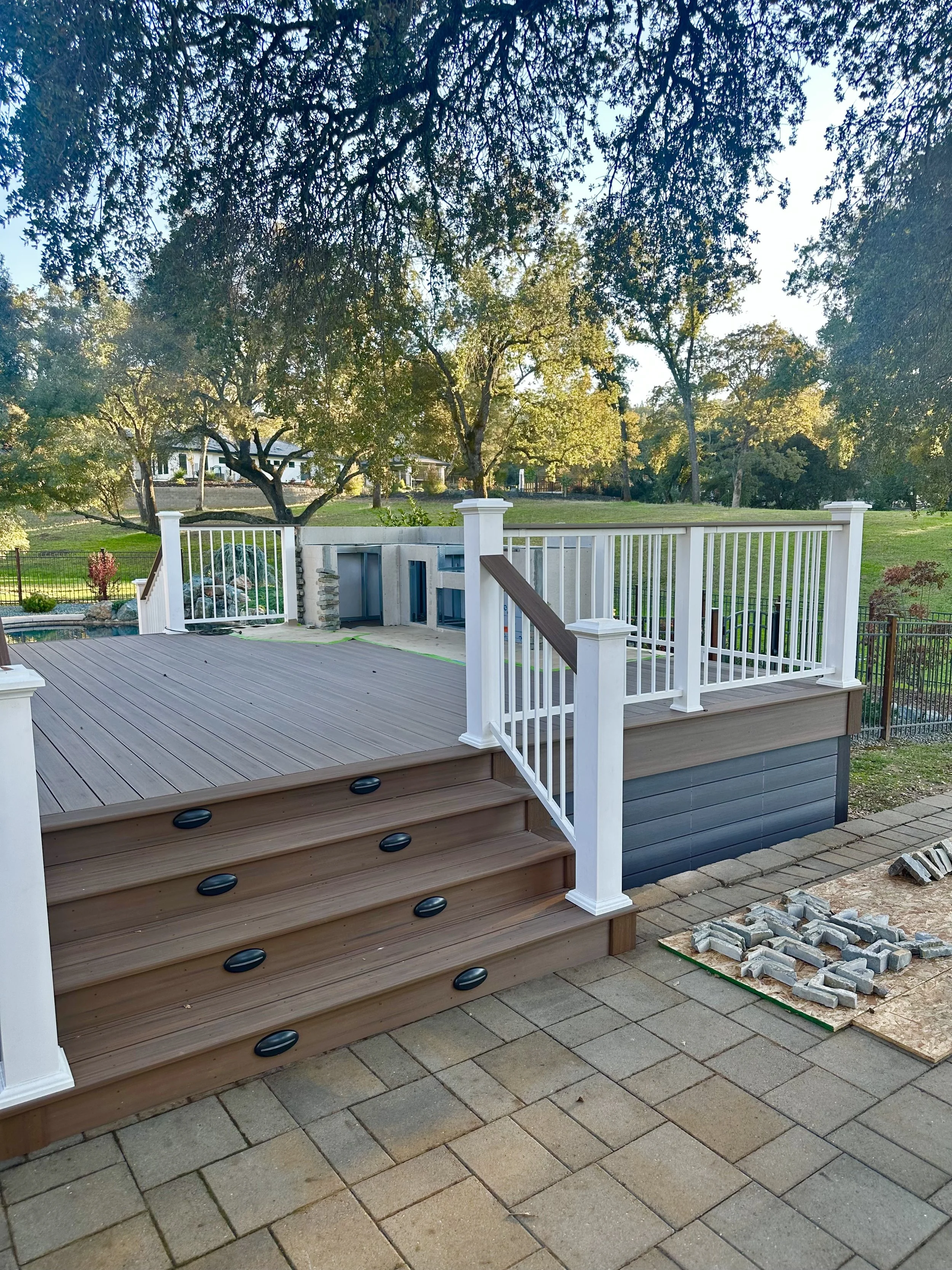 A wooden deck with stairs leading up to a backyard, surrounded by a white railing, with a landscaped yard and trees in the background.