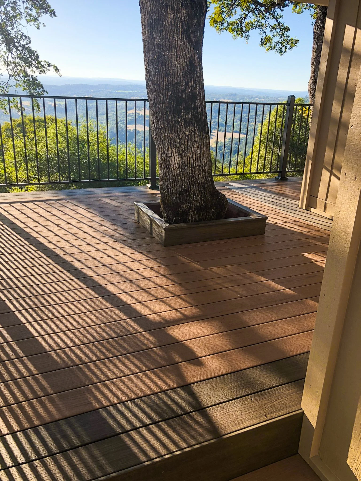 Wooden deck with a potted tree, metal railing, and a scenic view of hills and forests in the distance.