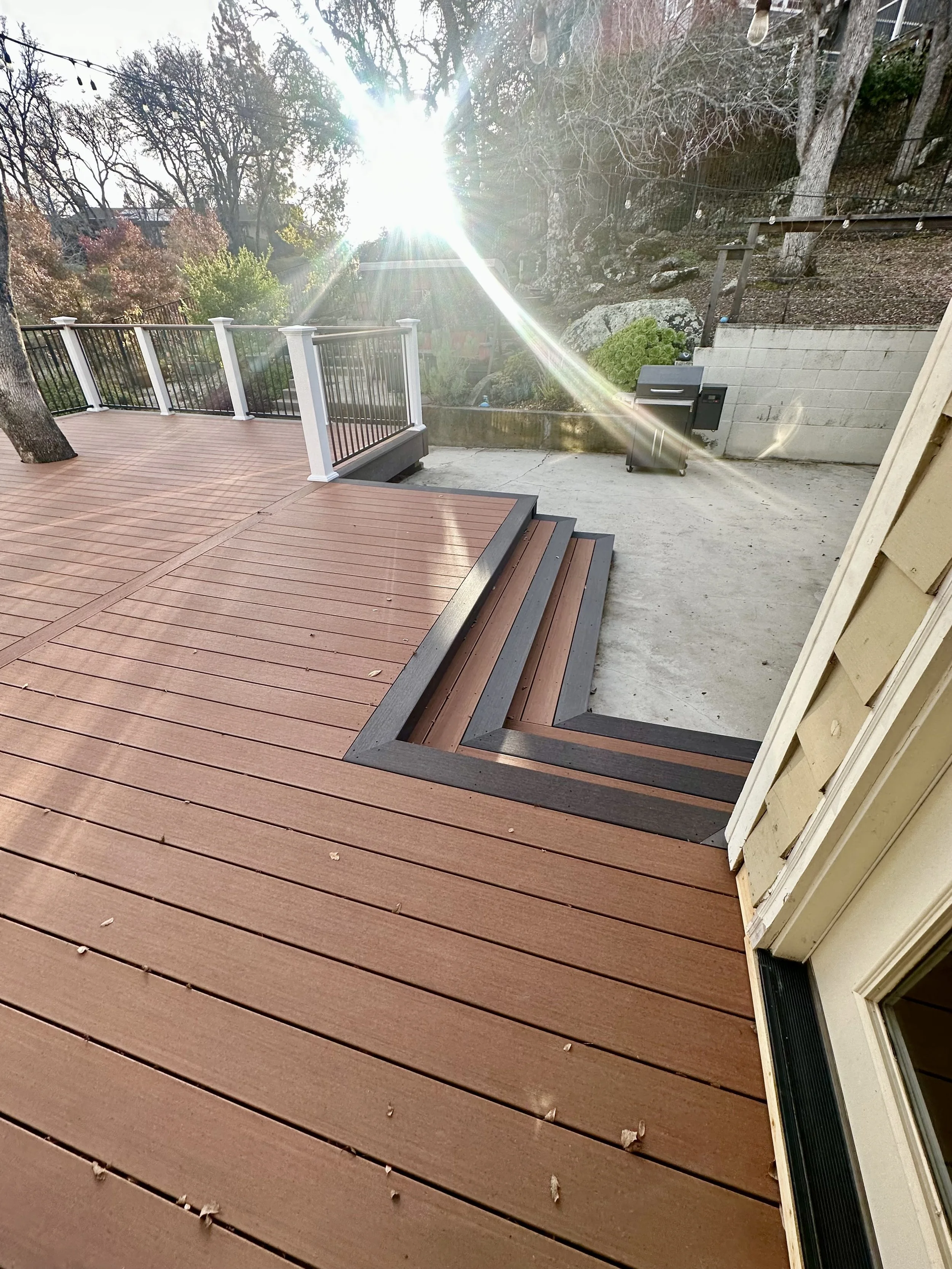 A wooden deck with stairs leading down to a concrete patio area in a backyard, with a grill and a view of trees and a fence in the background.