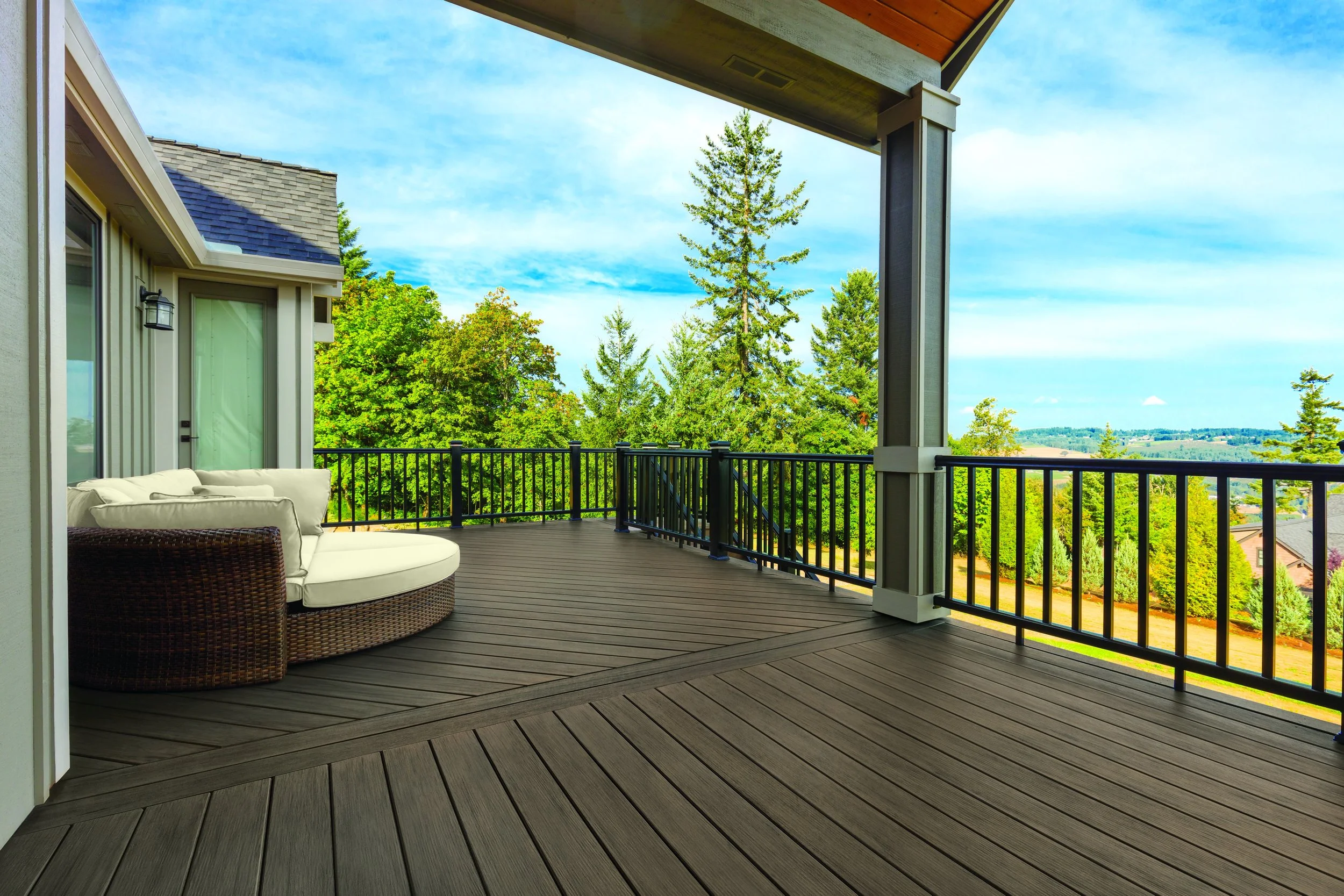 Empty outdoor balcony with wooden flooring, white cushioned outdoor furniture, black metal railing, overlooking green trees and a blue sky with clouds.