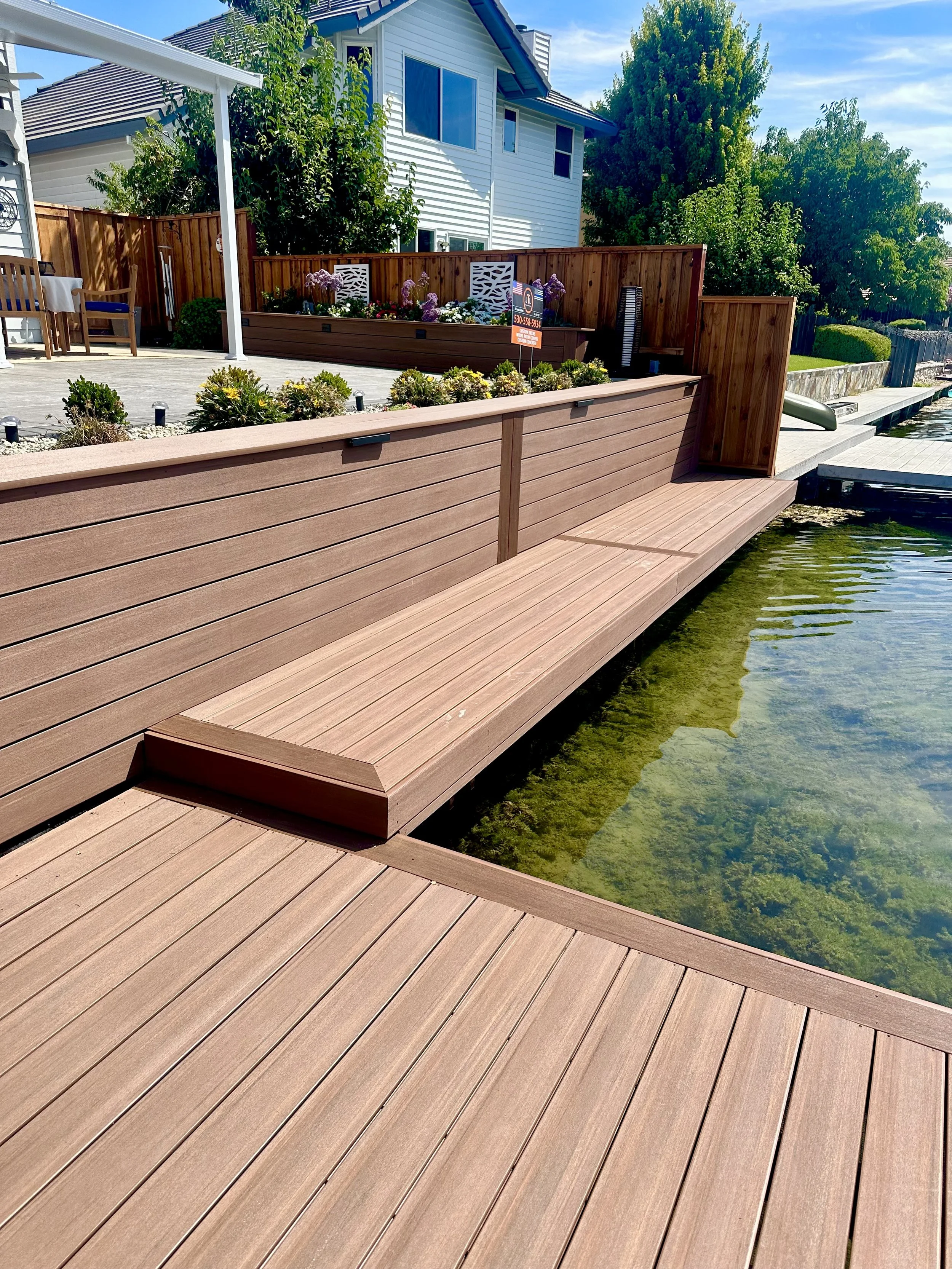 A wooden dock extending over a water body with clear water and some aquatic plants, adjacent to a backyard with a patio, plants, and a house in the background. The patio has outdoor furniture and a wooden privacy fence.