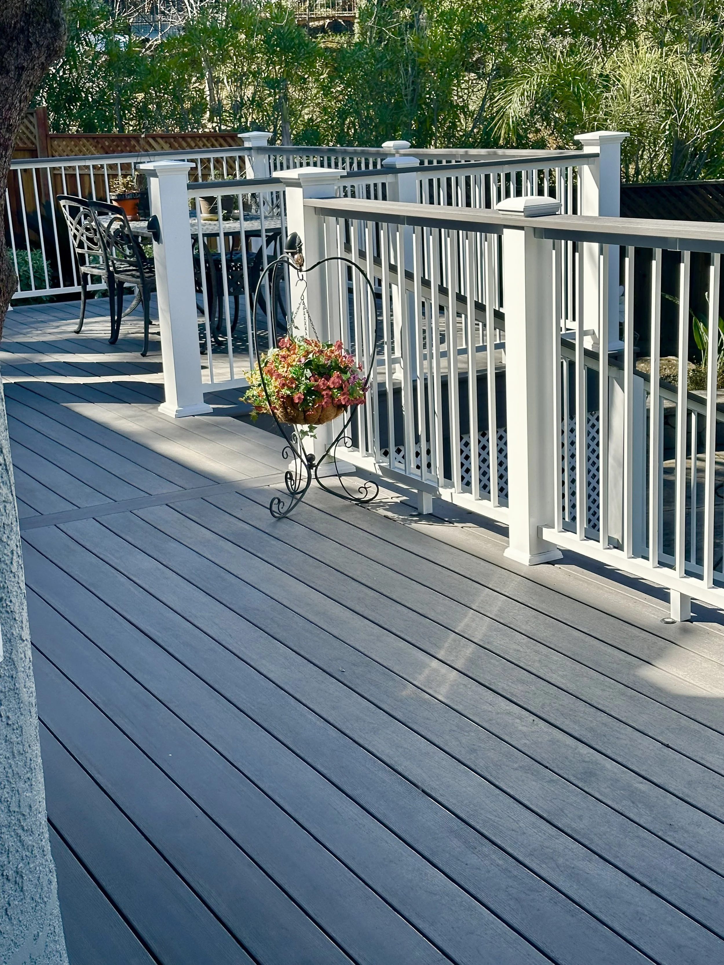 A wooden deck with white railing, a black metal outdoor dining table and chairs, a hanging flower basket, set in a backyard with lush greenery and trees.