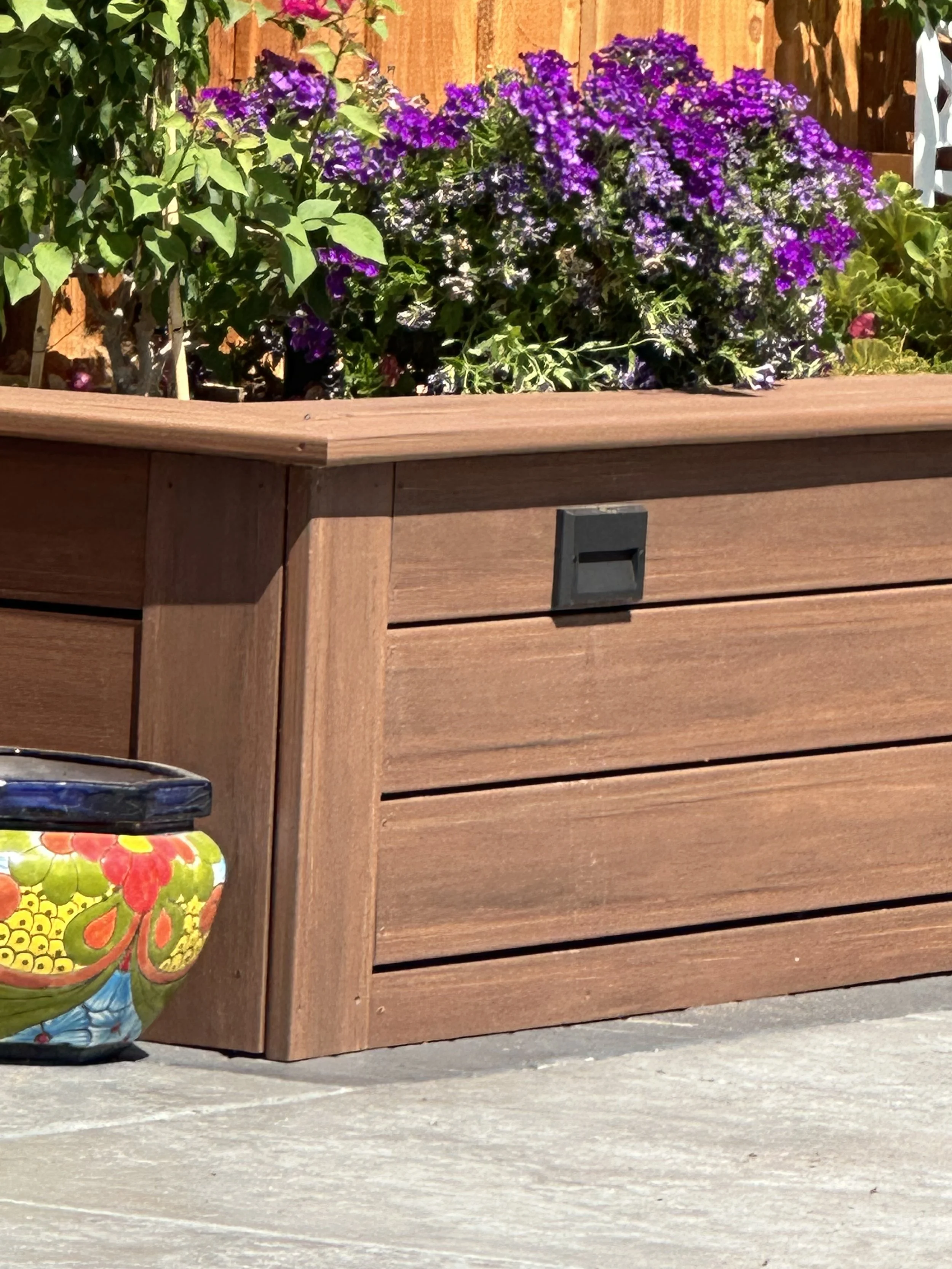 A wooden planter box with purple flowers, some green leaves, and a colorful ceramic pot on the ground beside it. The background has a wooden fence.