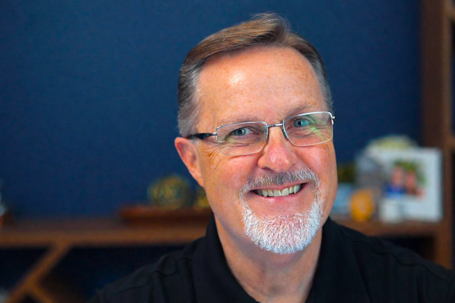 A middle-aged man with glasses, a beard, and a smile is wearing a blue polo shirt, sitting in a room with a bookshelf and personal items in the background.