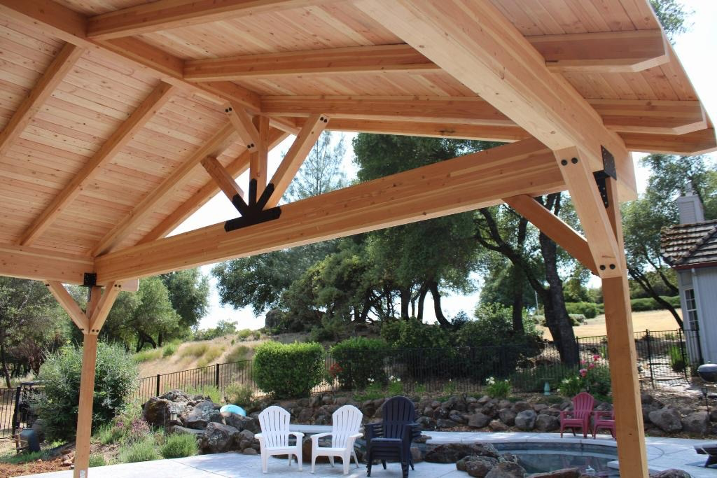 A wooden outdoor pavilion or gazebo with a sloped roof, situated in a backyard with trees, shrubs, and a stone-bordered garden. There are white and black chairs and a small table outside the pavilion.
