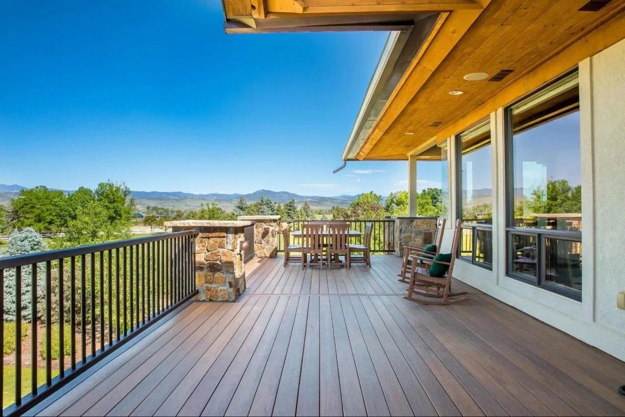 A spacious outdoor wooden deck attached to a house with large windows, surrounded by a black metal railing, overlooking a landscape with trees and mountains in the distance on a clear, sunny day.