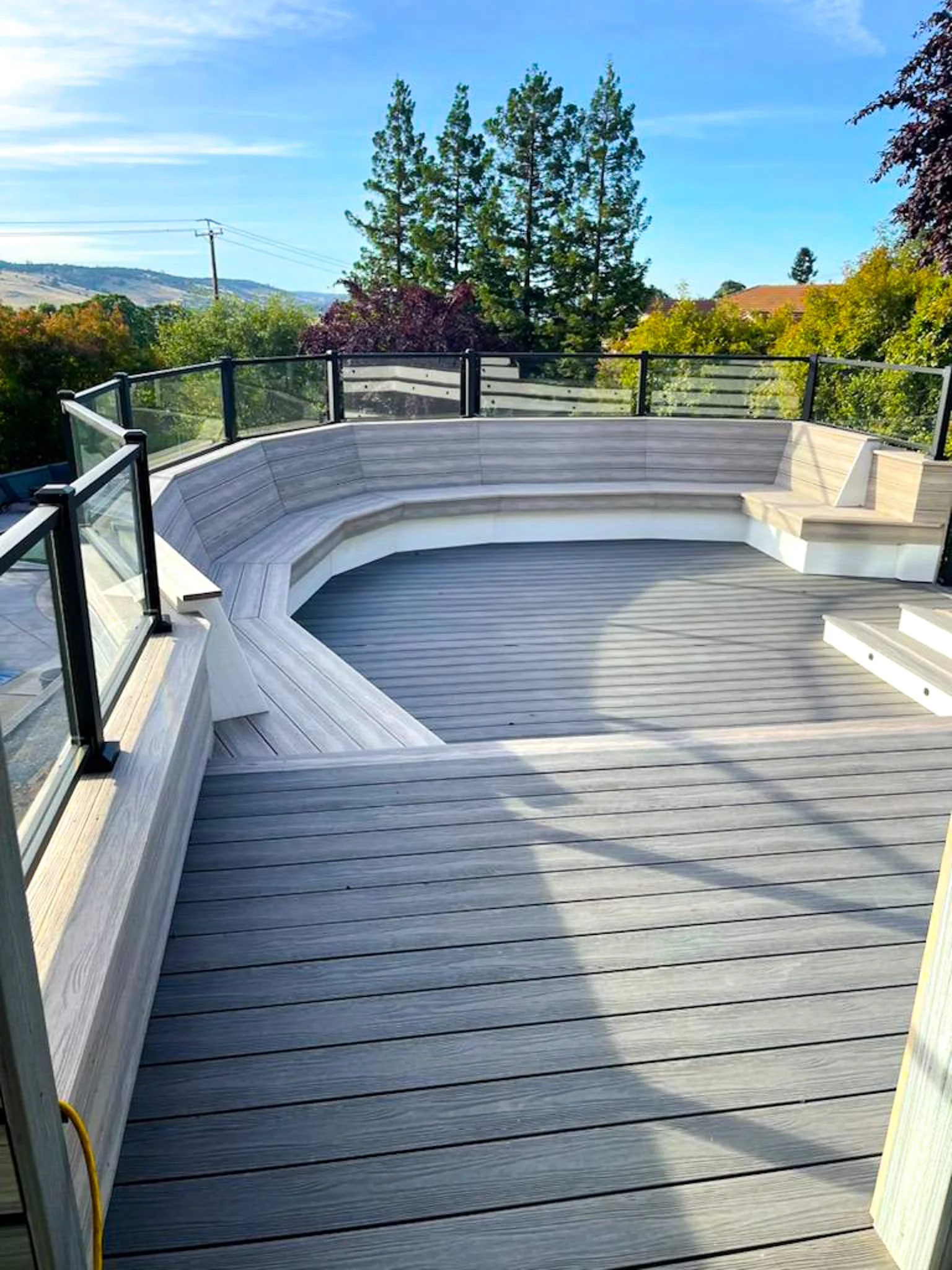 Empty outdoor wooden deck with glass and black metal railing, overlooking trees and hills under a clear blue sky.