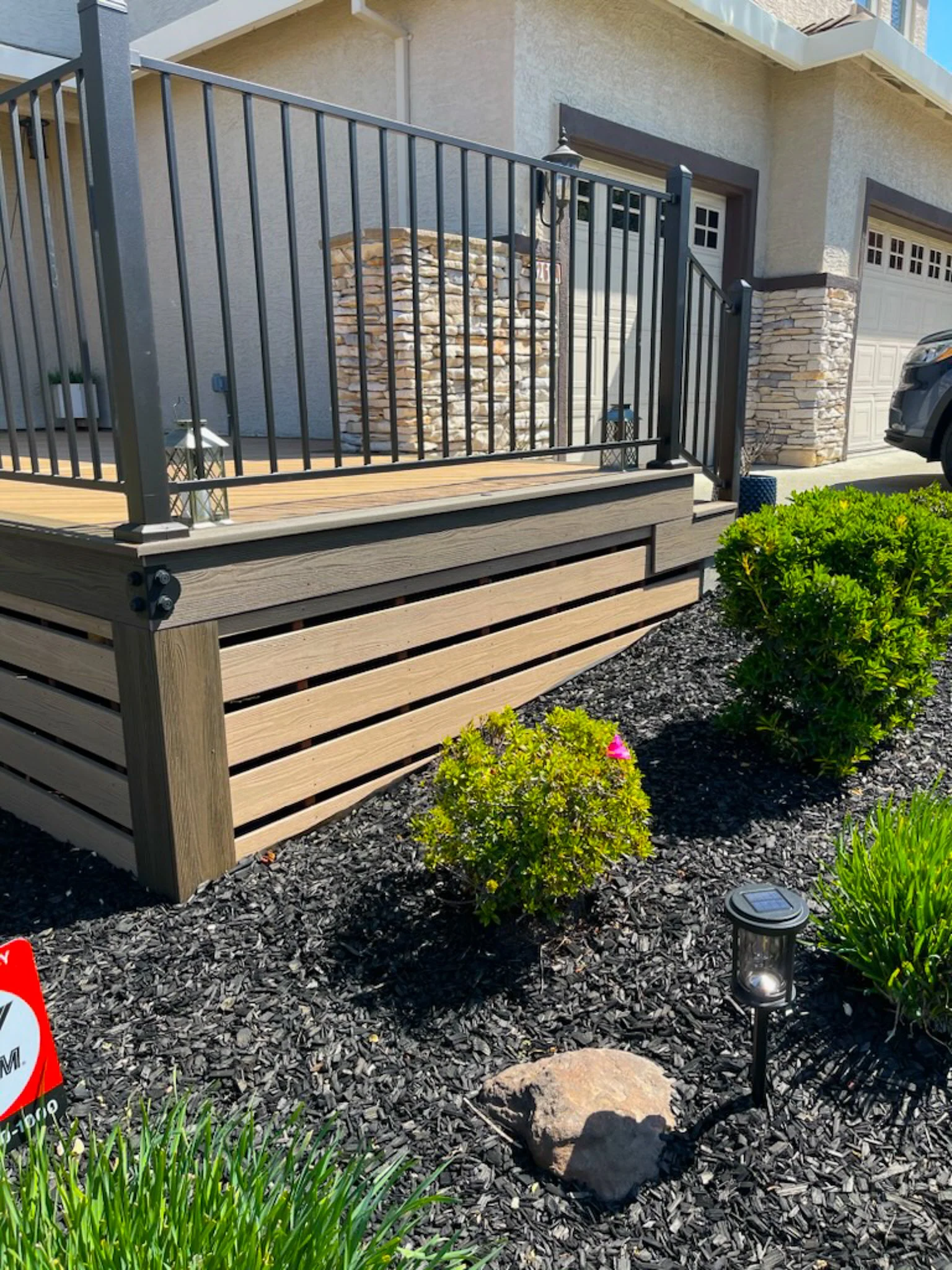 A residential front yard with a wooden porch bordered by a black metal railing, surrounded by landscaped garden beds with bushes, a large rock, and a solar-powered garden light.