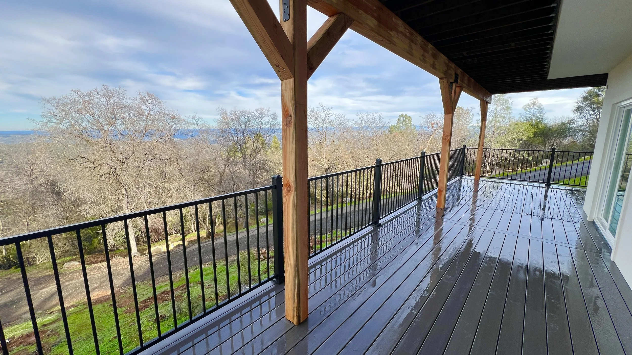 Rain-soaked wooden balcony with black metal railing overlooking a landscape of leafless trees and distant blue mountains under a cloudy sky.