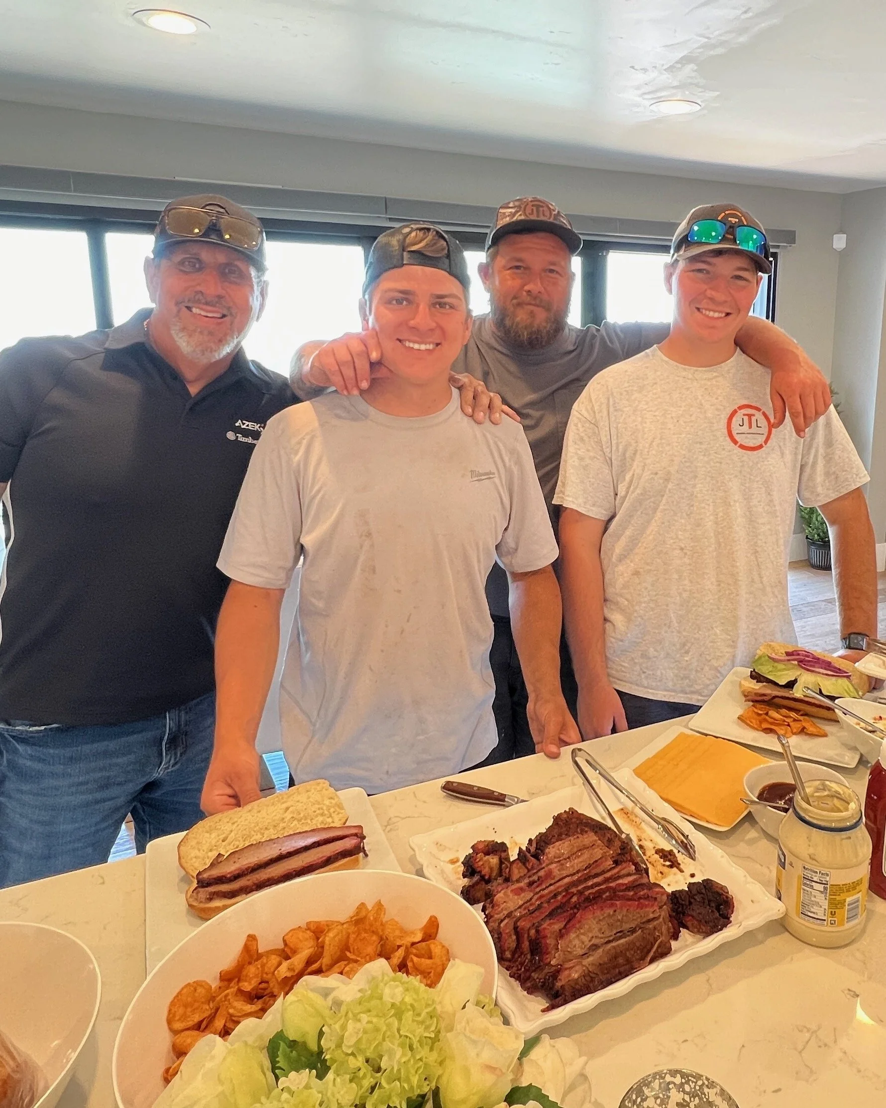 Four men stand behind a table with sliced brisket, a sandwich with meat and bread, and various side dishes, inside a well-lit room with a window.