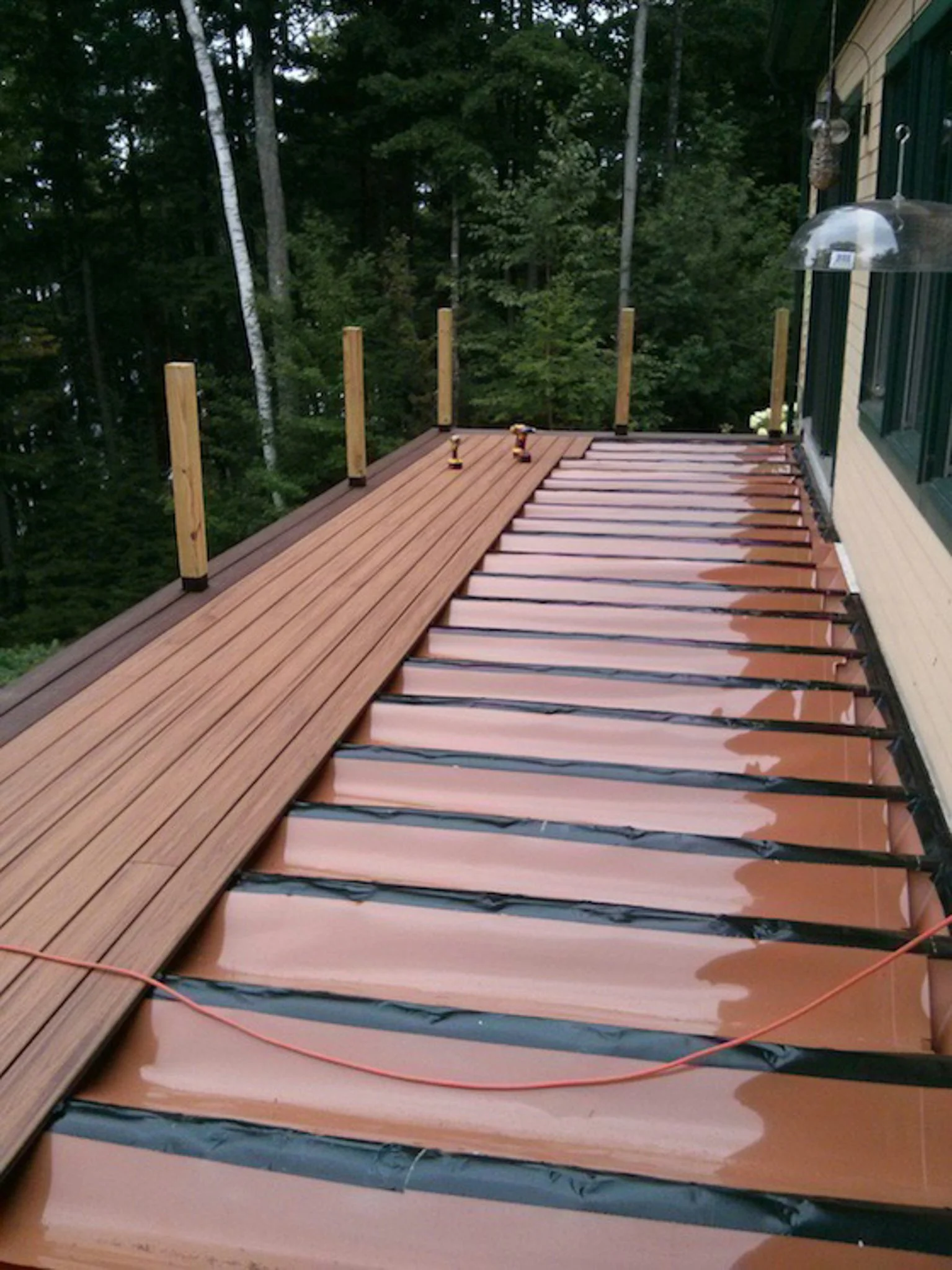 Wooden deck under construction on a house, with some decking boards installed and others still to be put down, surrounded by green trees.