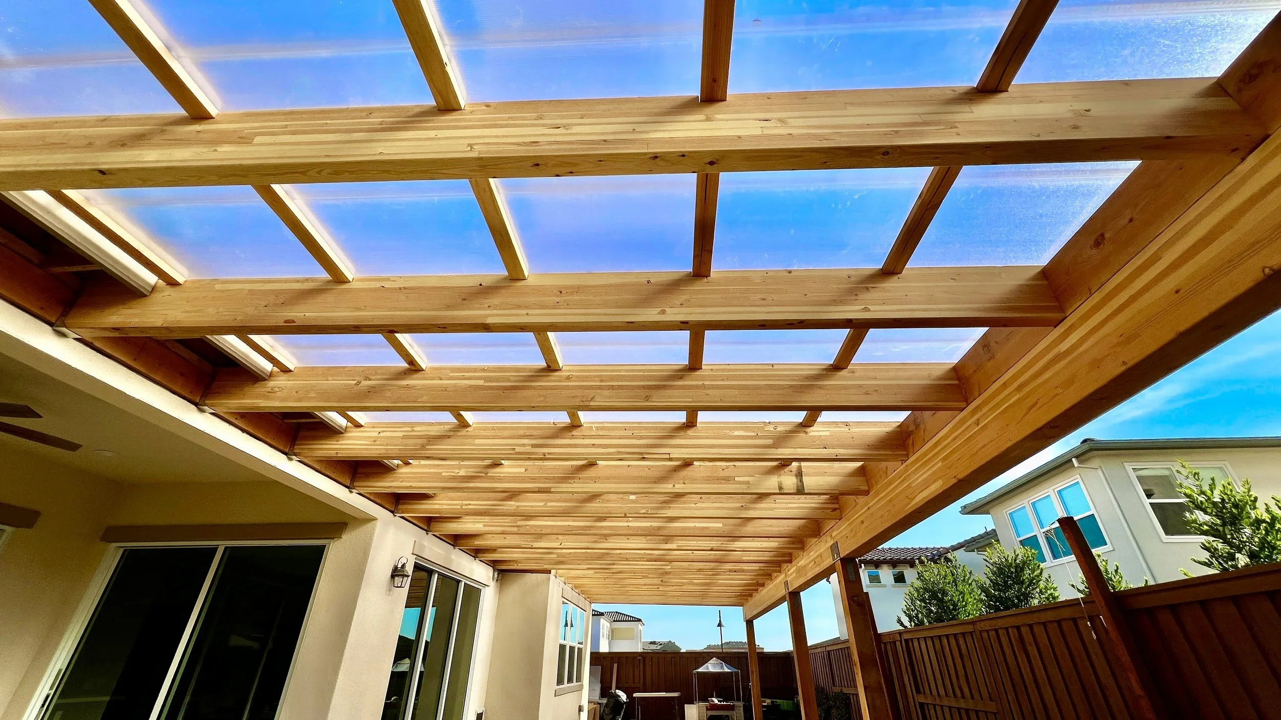 View of a newly constructed wooden pergola or patio cover attached to a house with clear blue sky above, outdoor furniture and greenery visible.