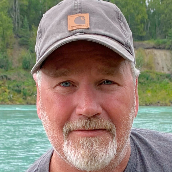 Close-up of a man with a beard and mustache wearing a gray Carhartt cap, outdoors near a body of water with green trees and cliffs in the background.