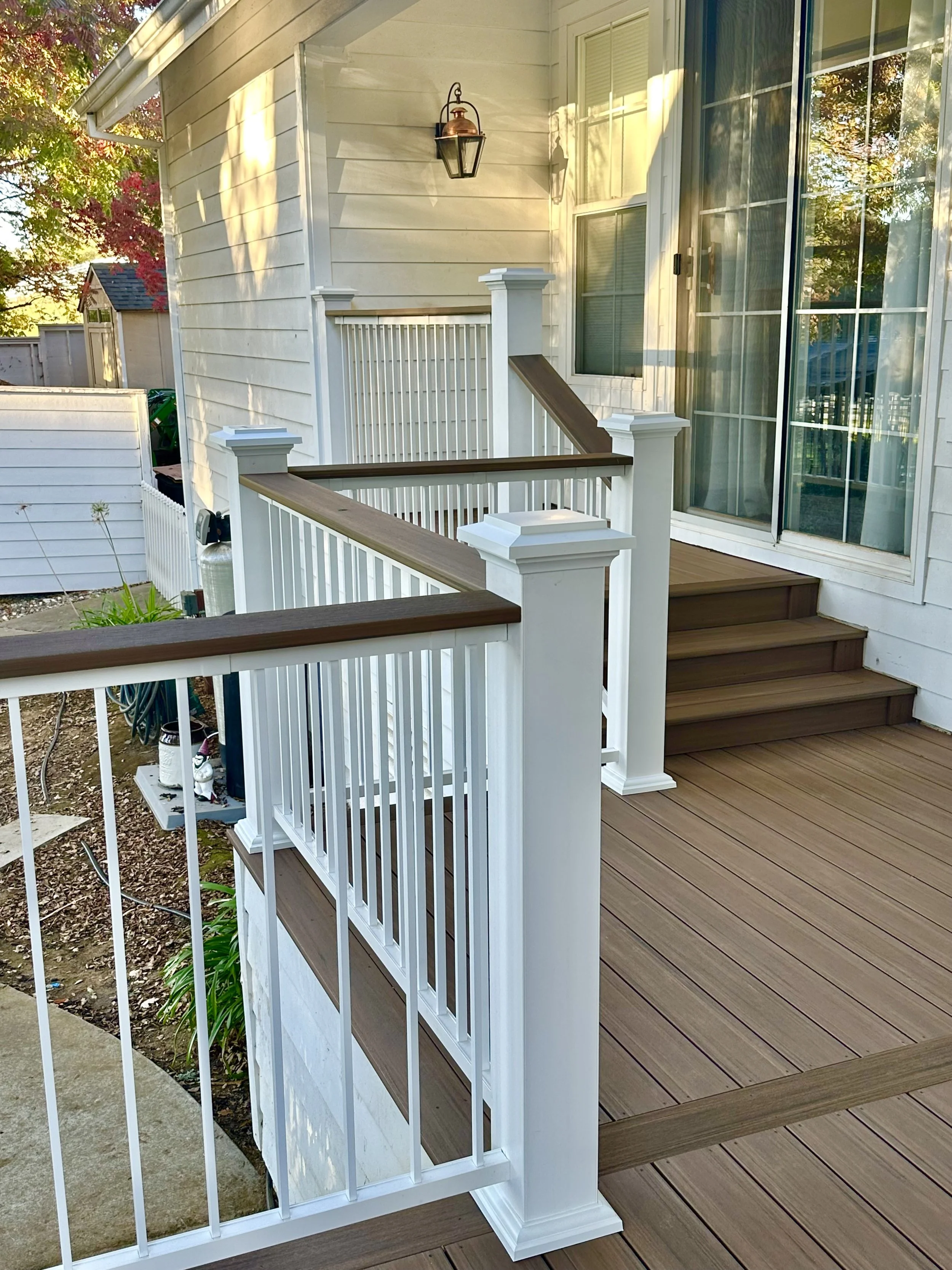 A wooden deck with white railings and brown handrails adjacent to a house with large windows. There are steps leading from the deck and a lantern-style light fixture on the house wall. The scene shows trees with colorful autumn leaves in the backgrou