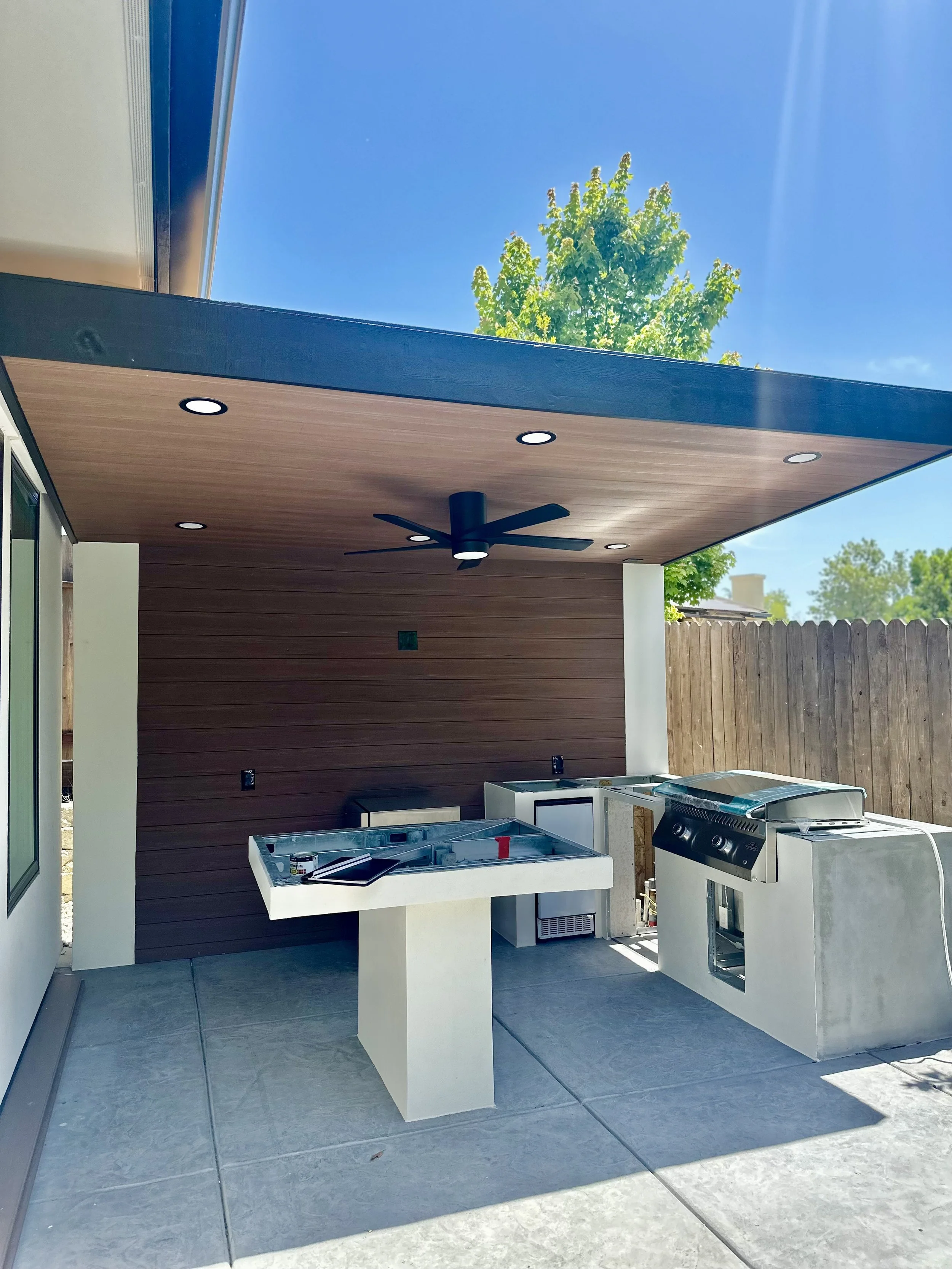 Outdoor patio area with a ceiling fan, recessed lighting, and a new outdoor kitchen setup including a grill, sink, and countertop, with a wooden fence and a tree visible in the background.