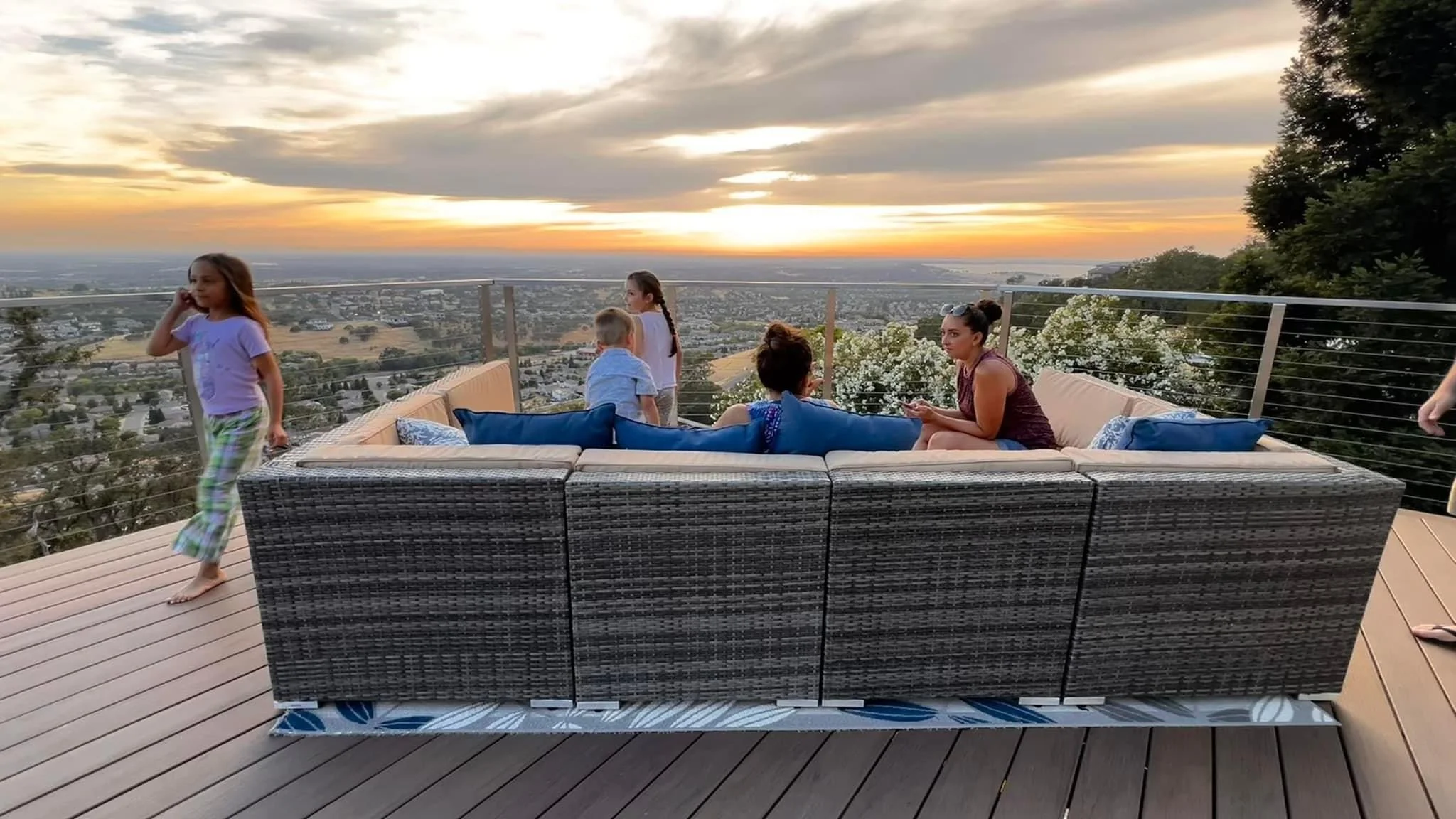 A group of people, including children and adults, sitting and standing on a rooftop deck with outdoor furniture by a glass railing, watching a sunset over a cityscape with trees and buildings.