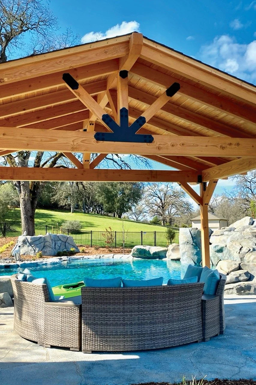 A backyard with a swimming pool surrounded by rocks, a black metal fence, and a grassy hill with trees. There is a wooden pavilion with a sloped roof overhead and outdoor seating with cushions. The sky is blue with white clouds.