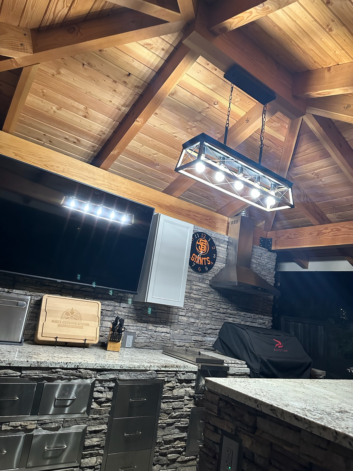Inside a rustic kitchen with a wooden vaulted ceiling, a black rectangular pendant light fixture hanging from chains, a large flat-screen TV mounted on a stone wall, a Golden State Giants clock, a charcoal grill covered with a black cover, and a gran