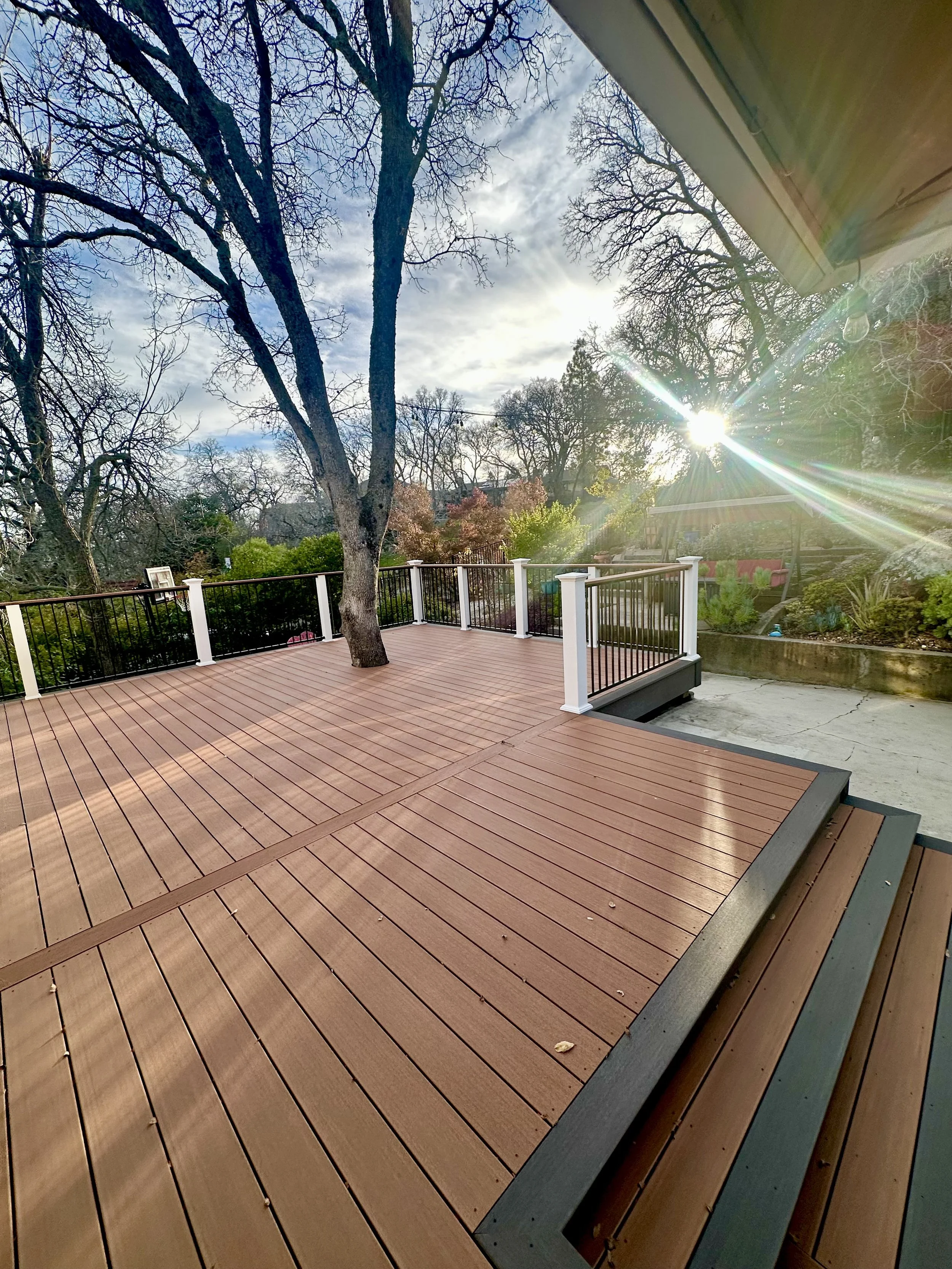 A backyard deck made of brown composite wood with black and white railing, trees in the background, and the sun shining through the sky.