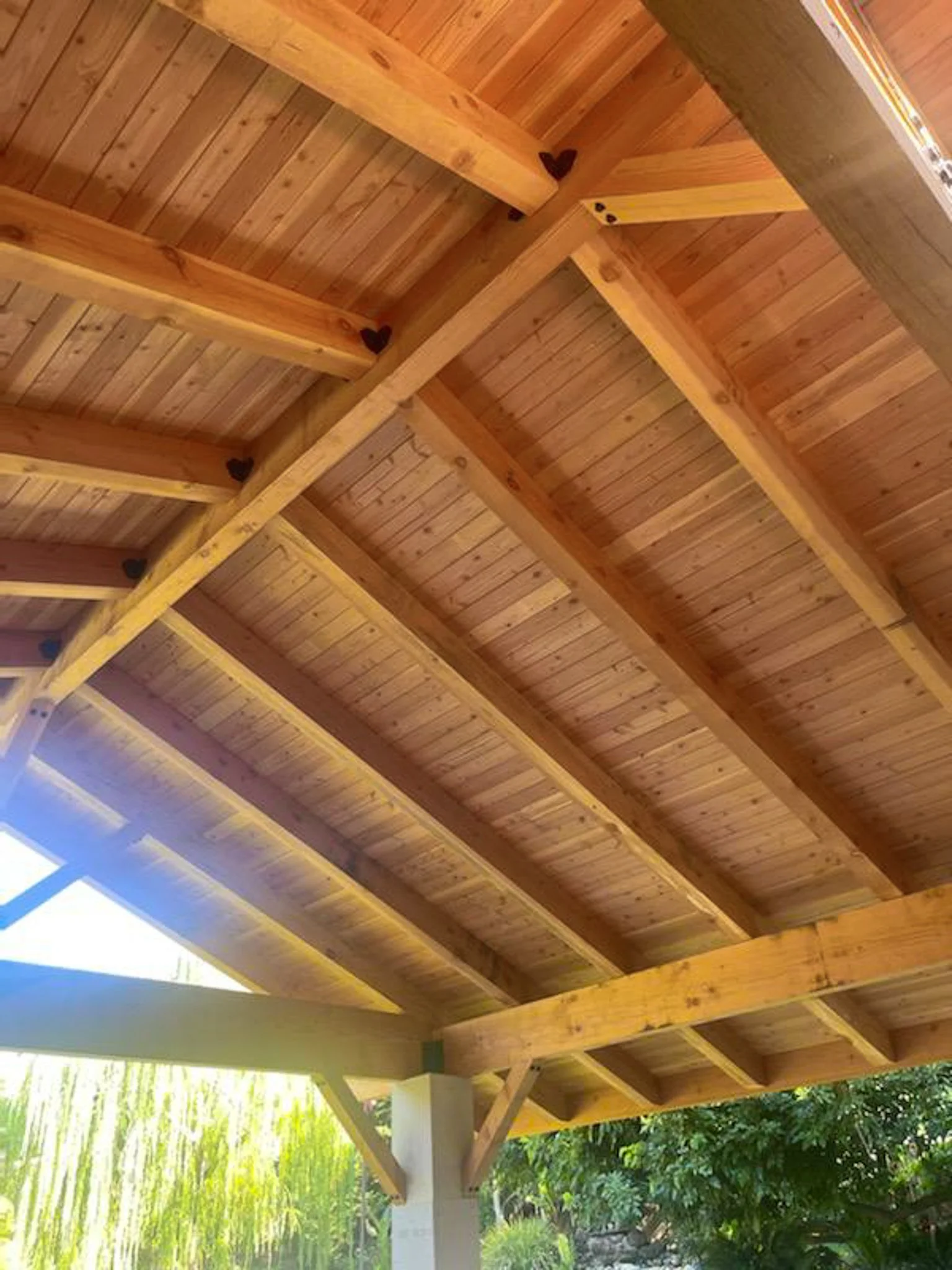 View of a wooden roof structure with exposed beams and slats, with some small decorative black hearts attached at the beams, and part of a garden with green trees and plants visible below.