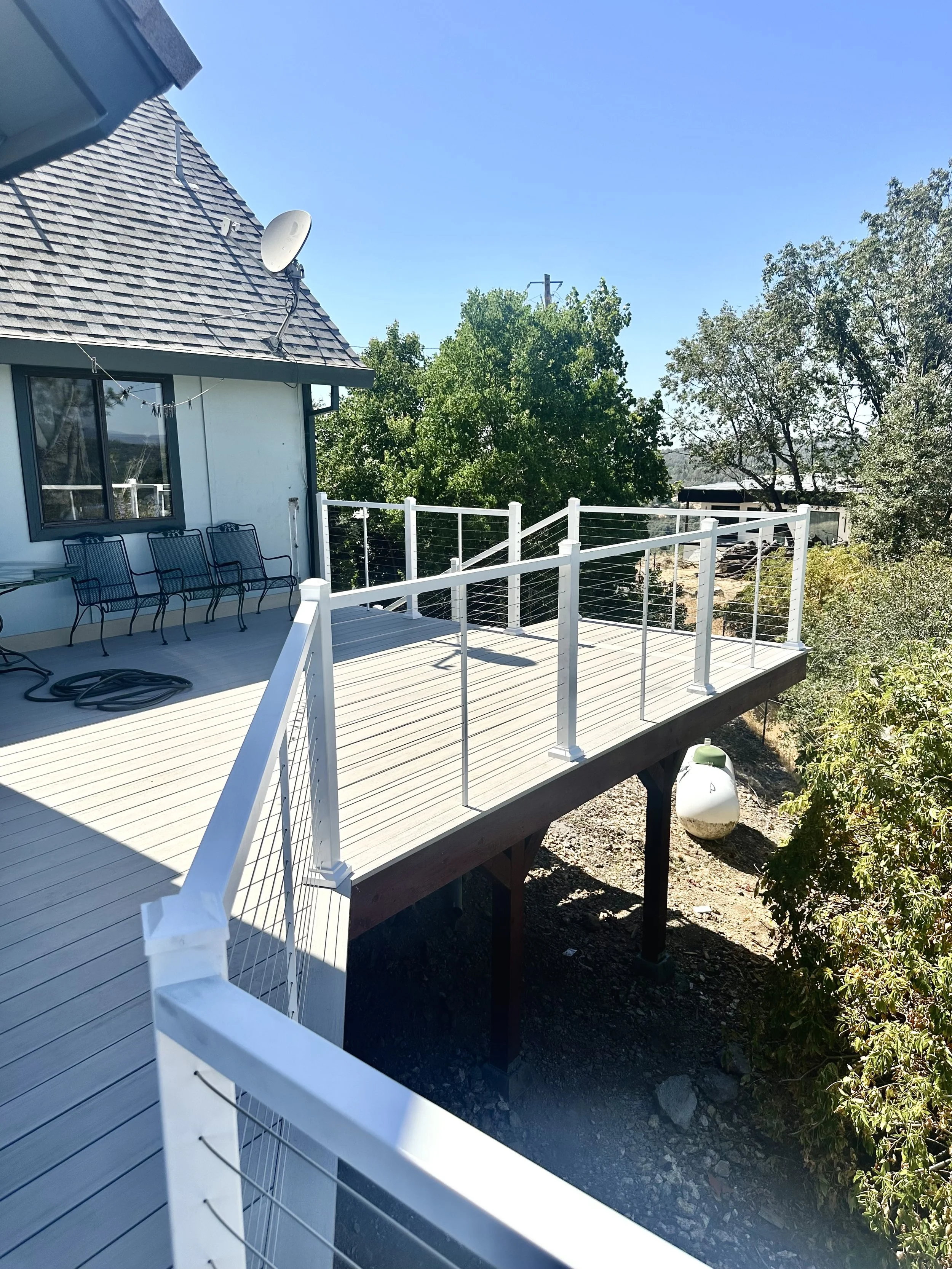 View of a wooden deck with a white railing extending from a house, overlooking trees and a gravel area below on a sunny day.