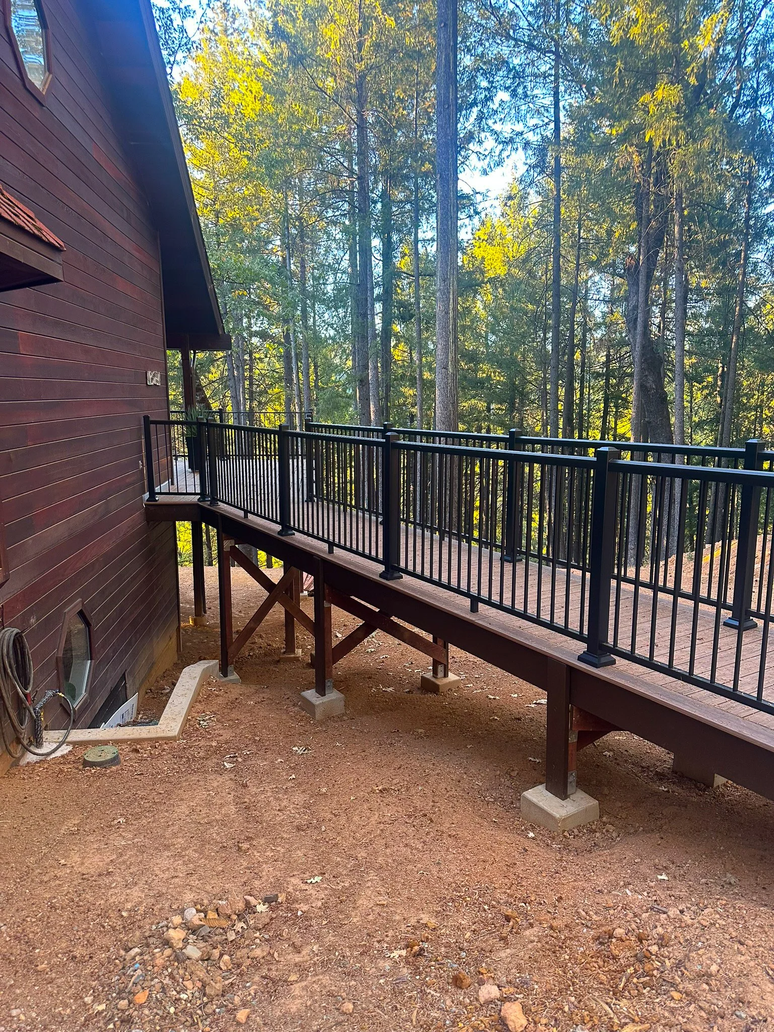 Wooden deck with black metal railing attached to a brown house, surrounded by tall trees and a forest in the background.