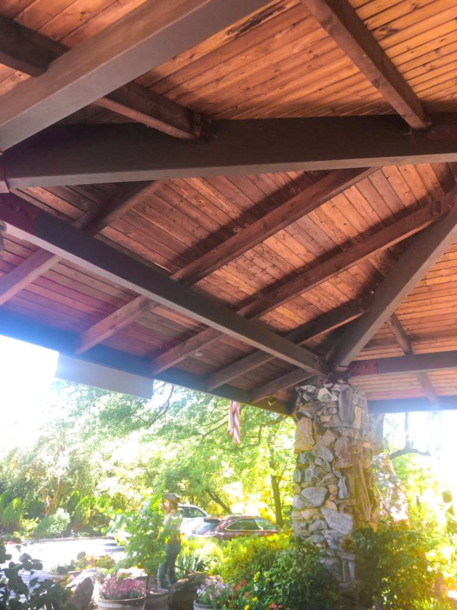 View of a wooden porch roof with wood beams and a stone pillar, surrounded by greenery and parked cars.