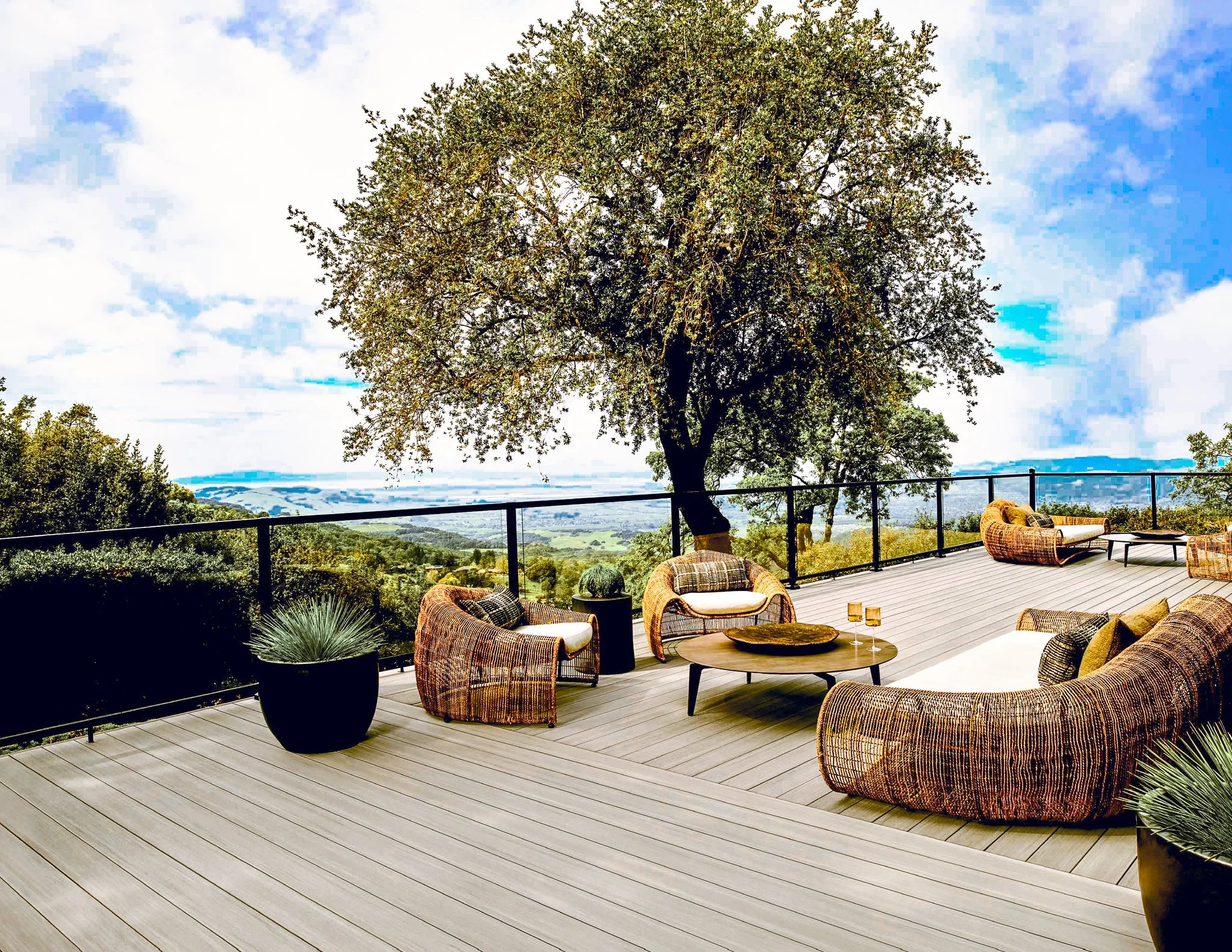 Outdoor deck with wicker furniture, including chairs and a sofa, overlooking a scenic landscape with hills and a large tree under a partly cloudy sky.