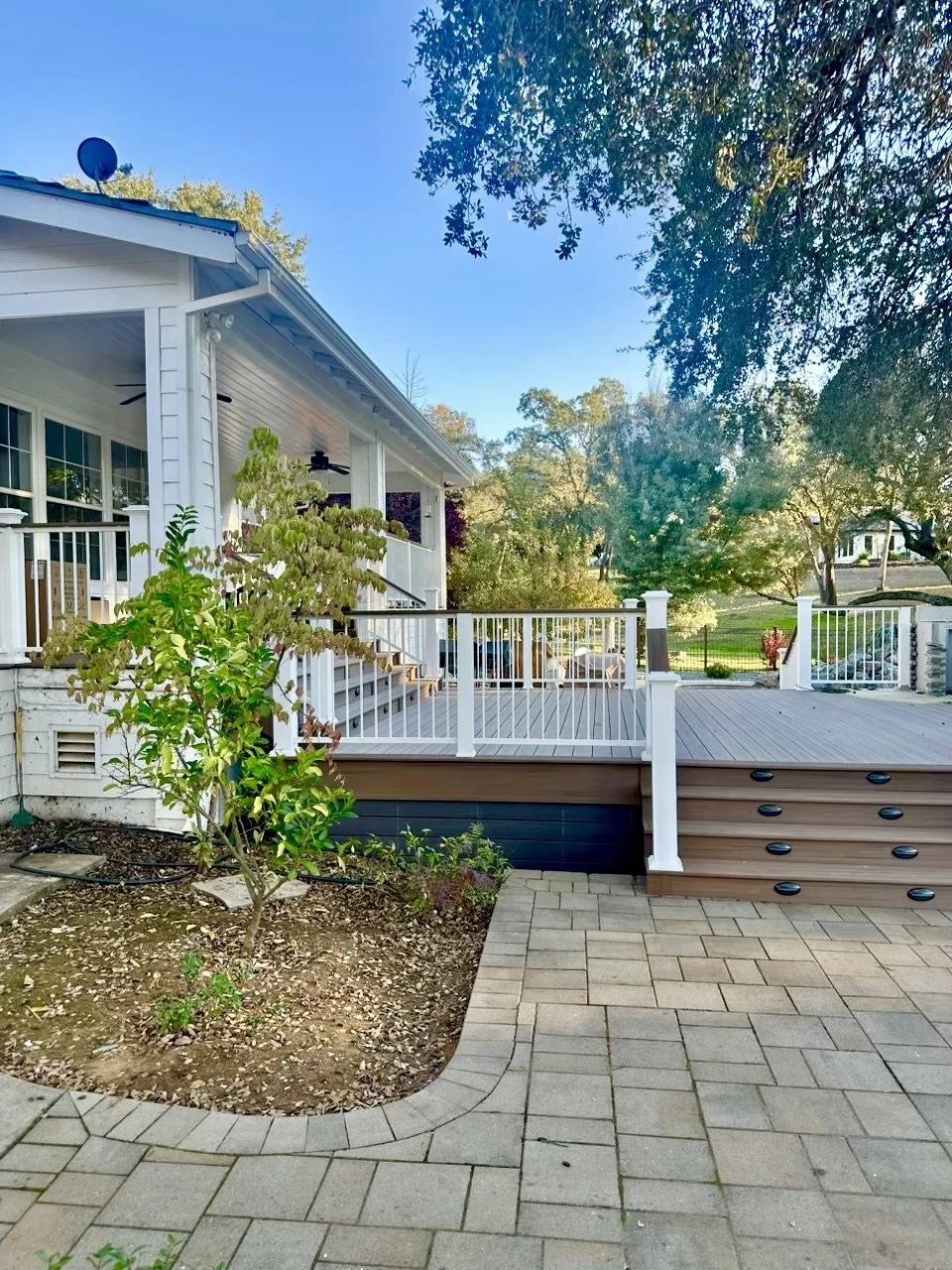A backyard patio with a raised wooden deck, white railing, steps, and a garden bed with plants and mulch. There are trees and a house in the background under a clear blue sky.