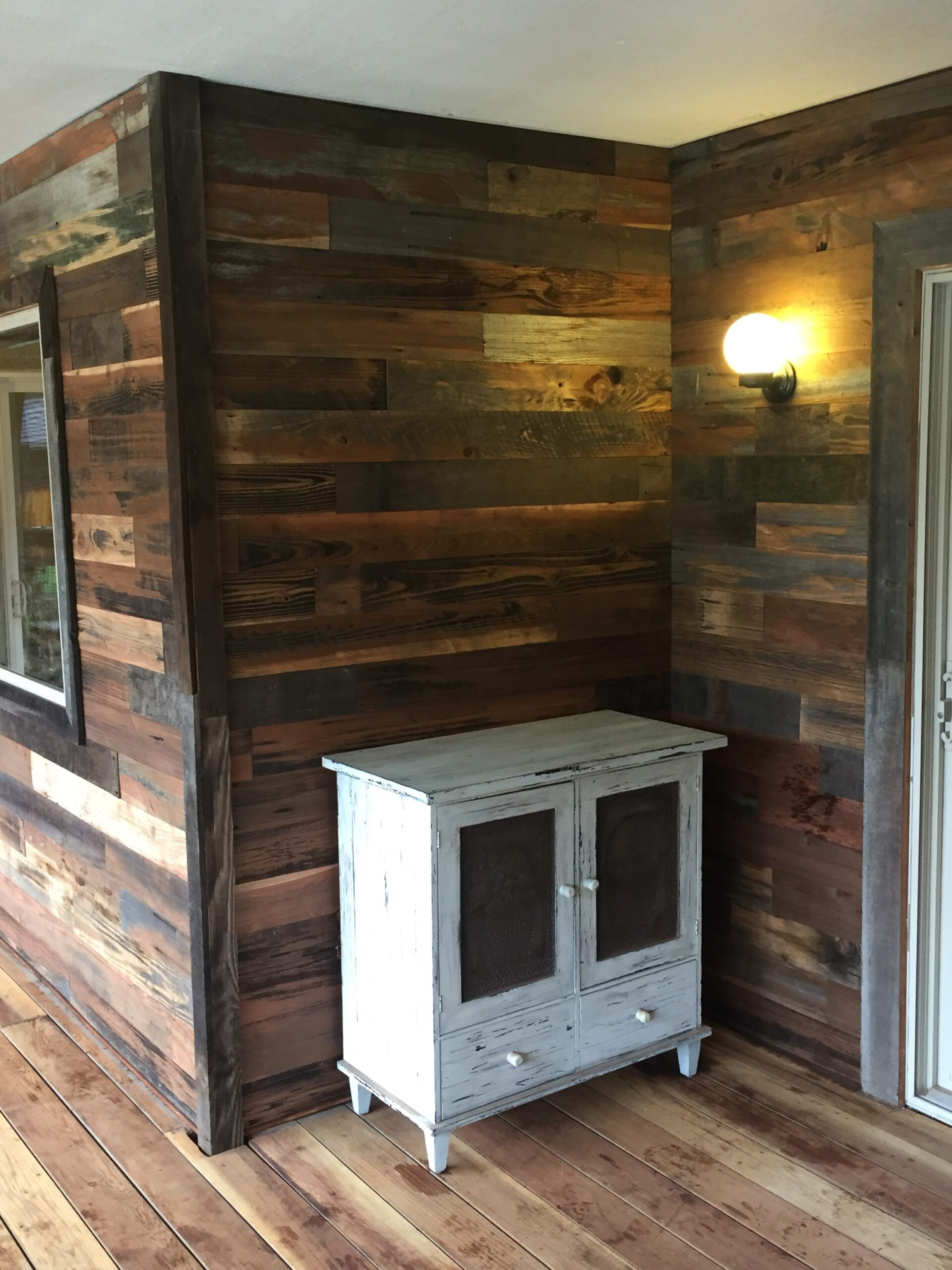 A rustic outdoor patio corner with a wooden wall, a small white distressed cabinet with mesh doors, and a wall-mounted light fixture illuminating the space.