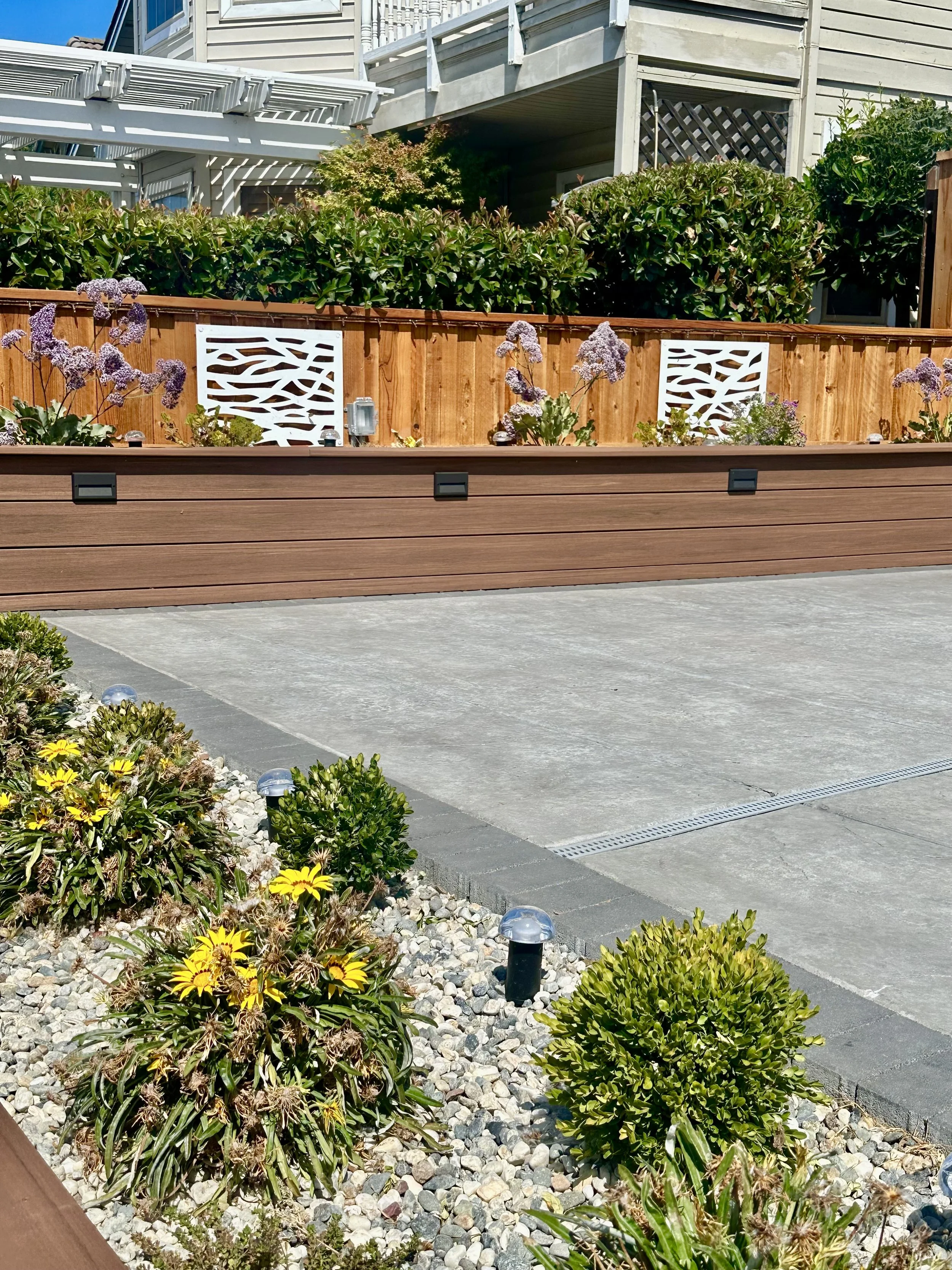 A backyard with flower beds, bushes, and outdoor lighting, featuring a wooden privacy fence and a concrete patio area.