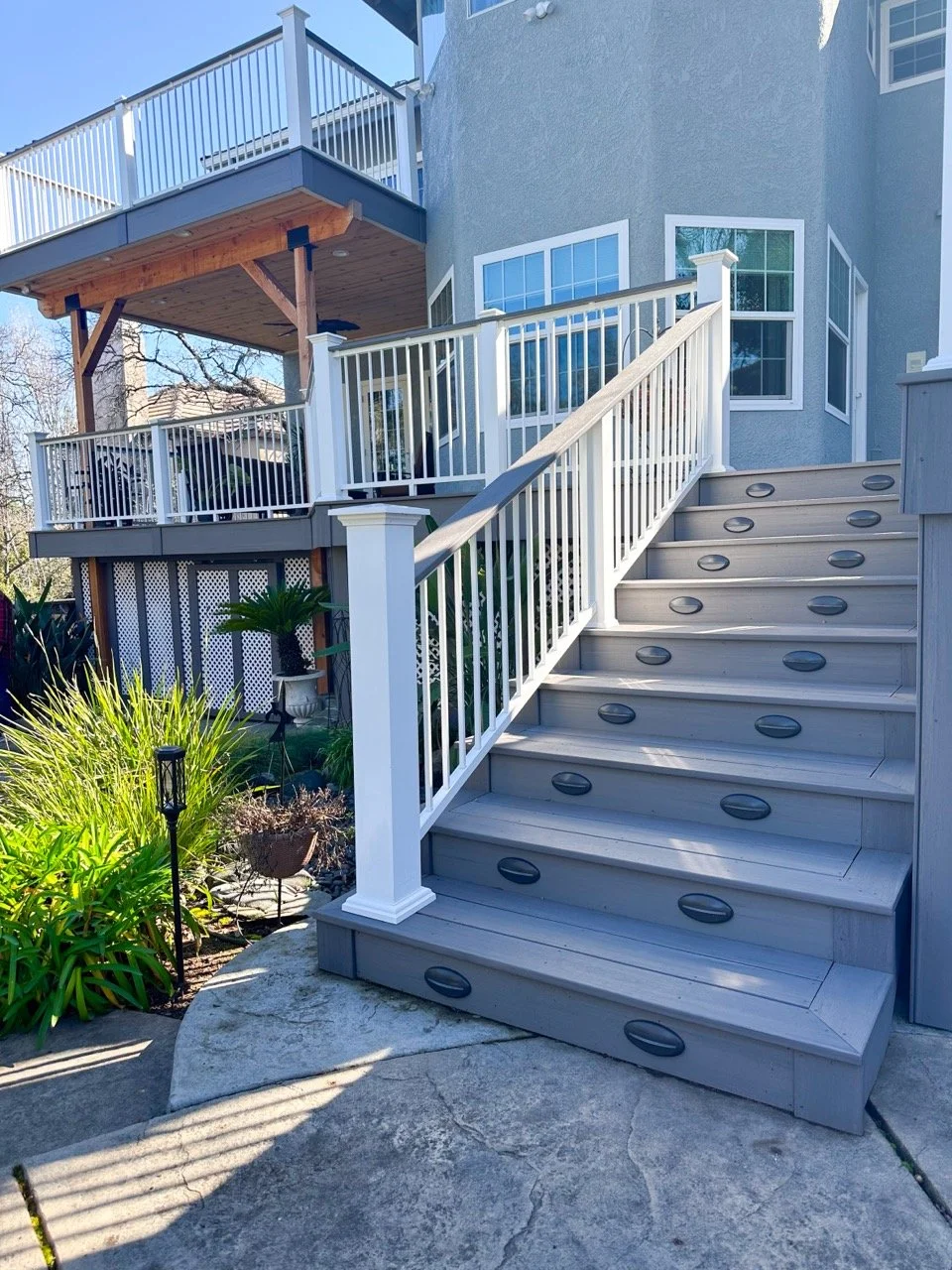 Outdoor wooden staircase leading up to a house deck with white railing, surrounded by plants and garden lights.