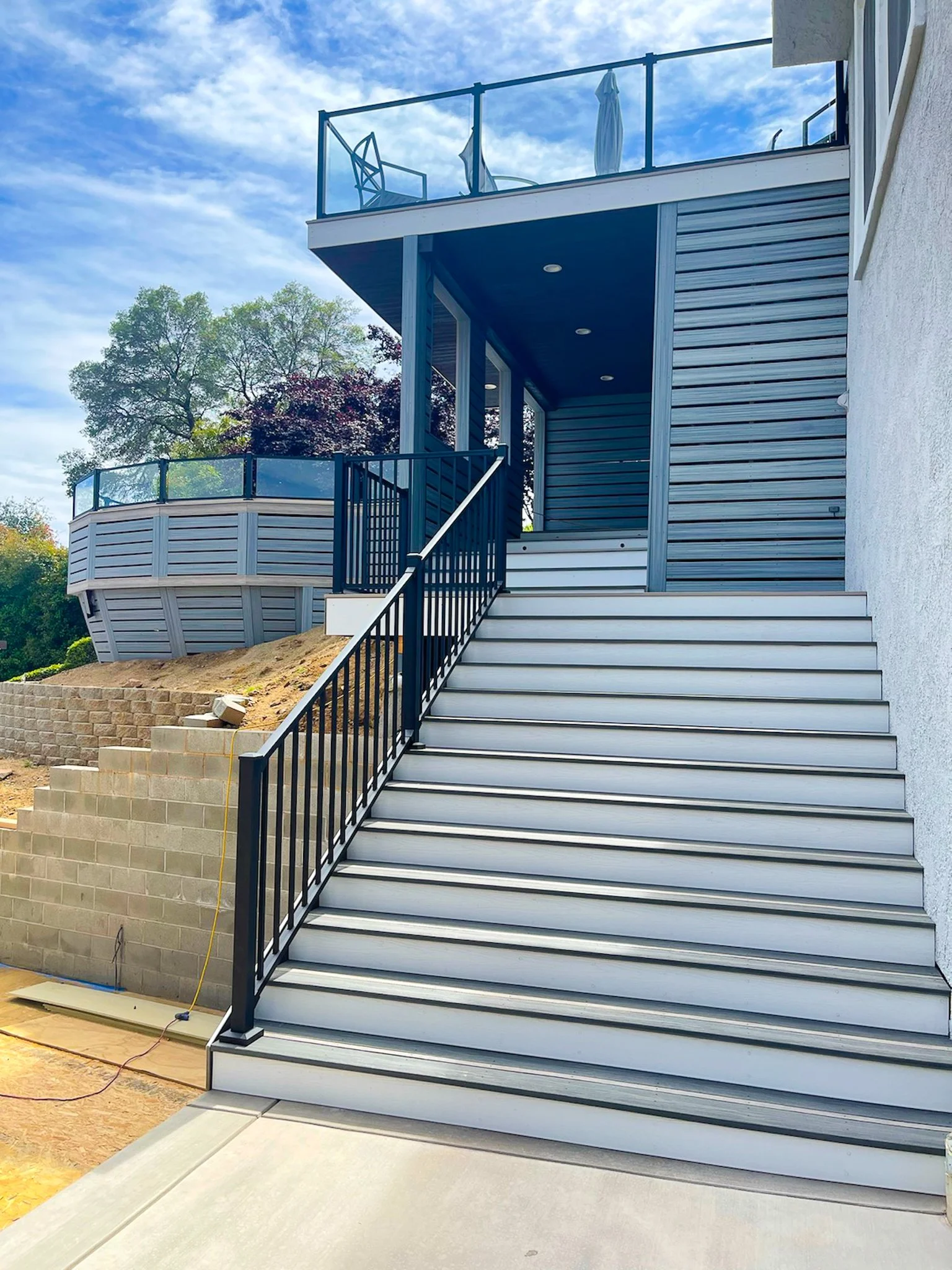 Exterior view of a modern house with a wide outdoor staircase leading to a balcony, with glass railings and patio furniture visible on the upper deck.