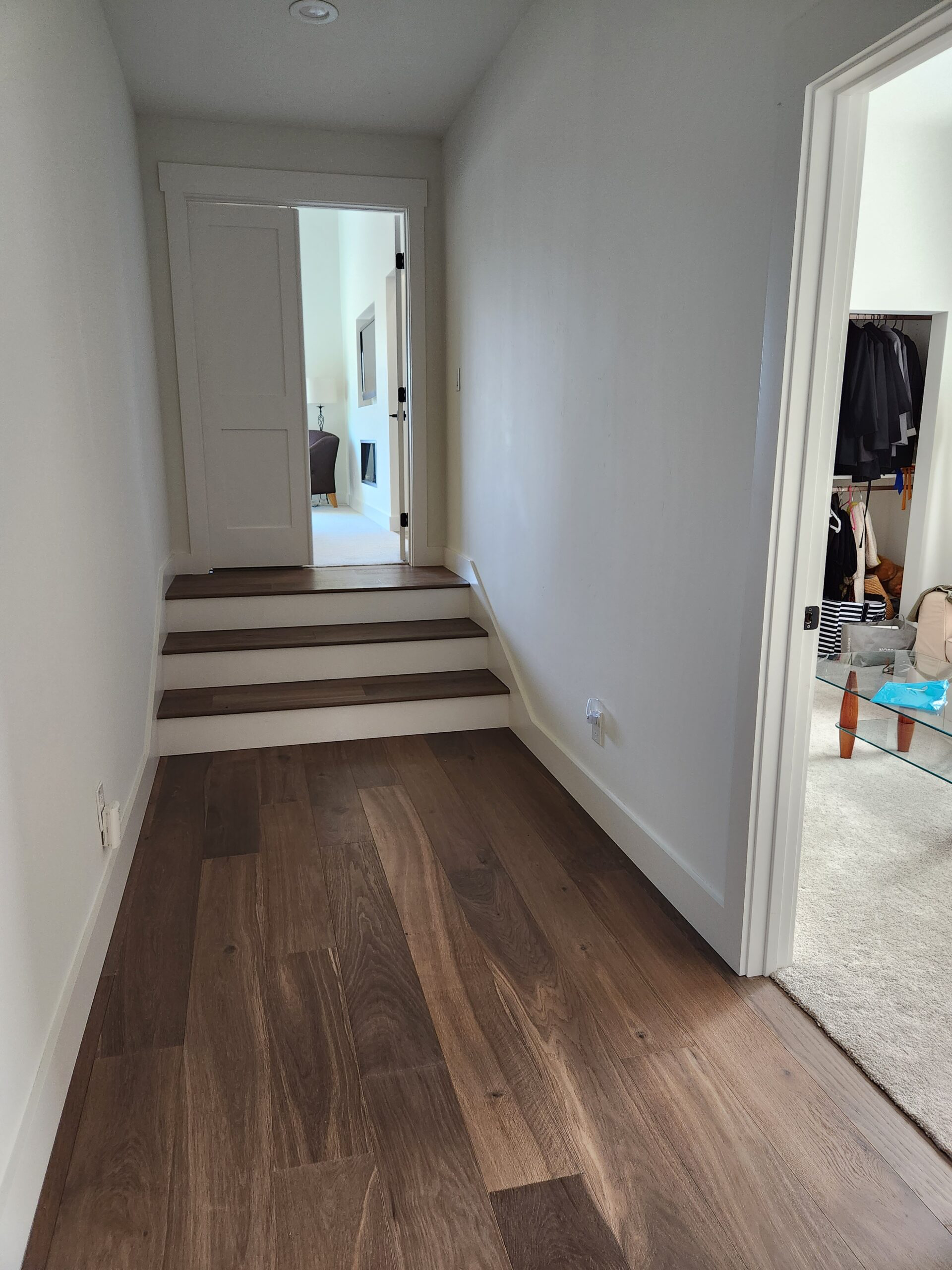 A hallway with wood flooring, leading to a staircase with three steps, and a door at the top of the stairs. To the right is an open doorway revealing a closet with hanging clothes and some storage boxes.