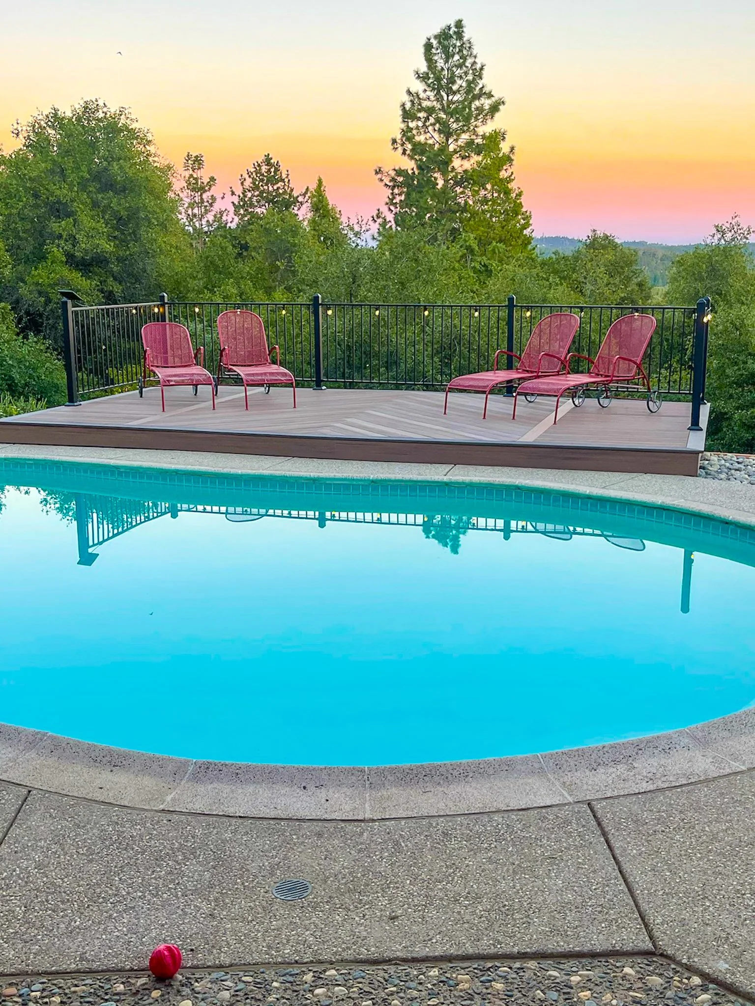 Poolside view of a backyard pool with a wooden deck and red lounge chairs, overlooking a green tree-filled landscape at sunset.