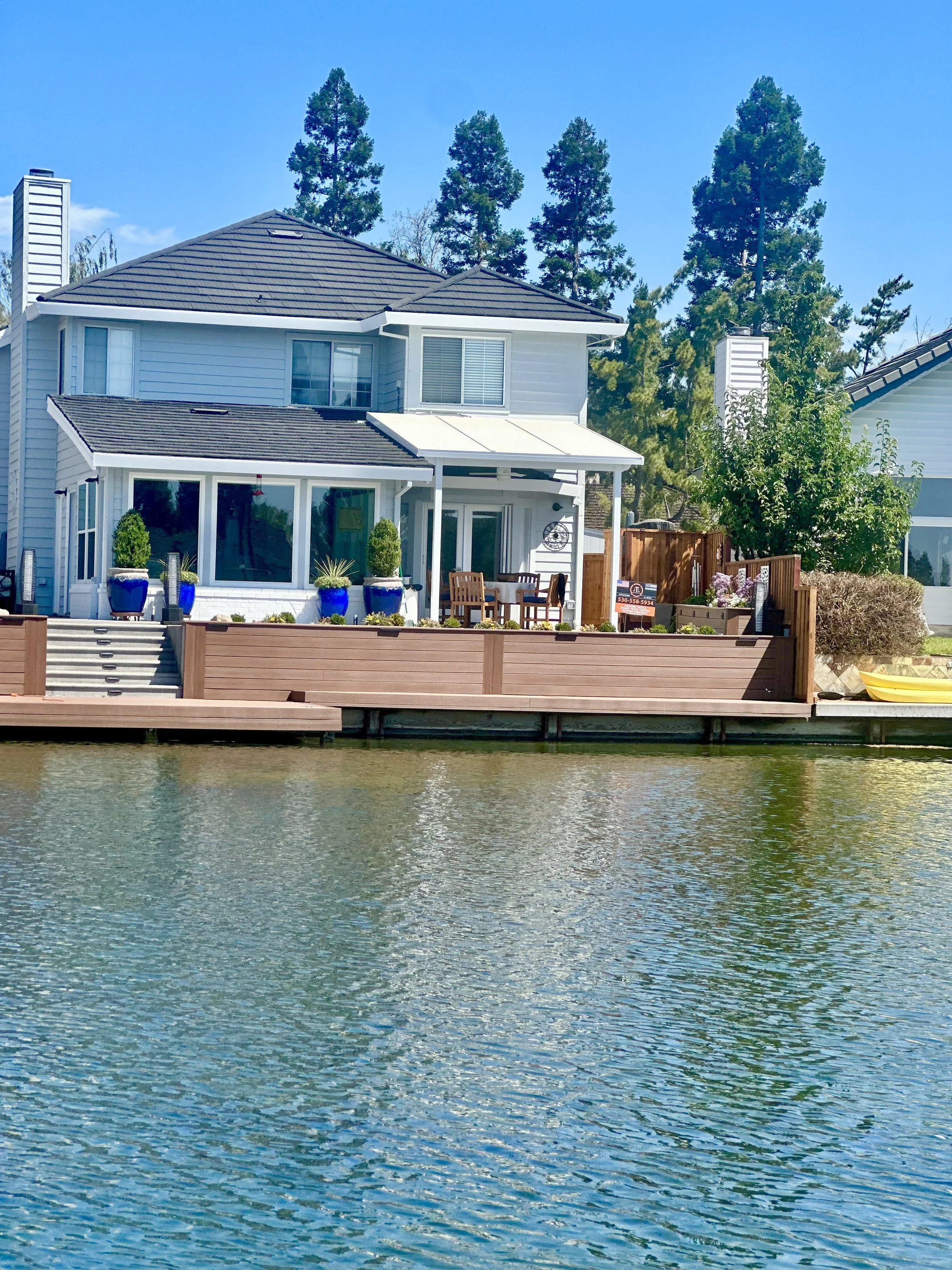  waterfront house with a deck, potted plants, and outdoor furniture, next to water with trees in the background.