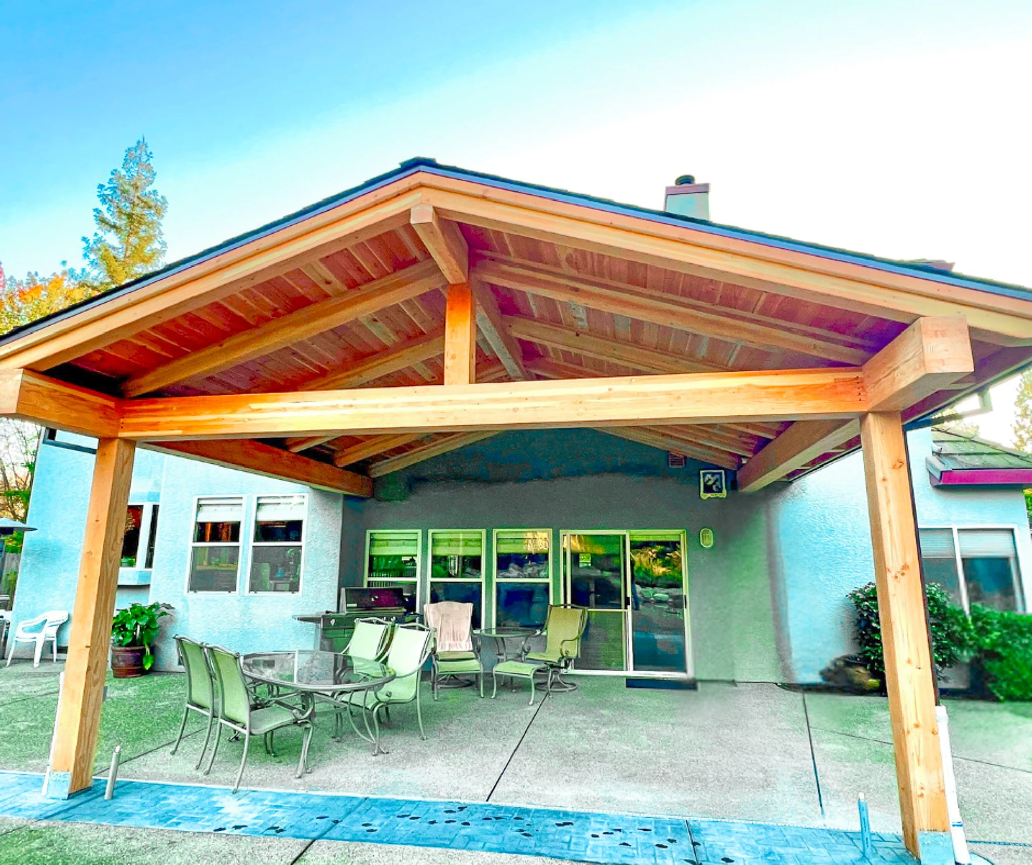 Backyard patio of a house with a newly constructed wooden pergola, outdoor furniture including a glass table and chairs, and a blue sky in the background.