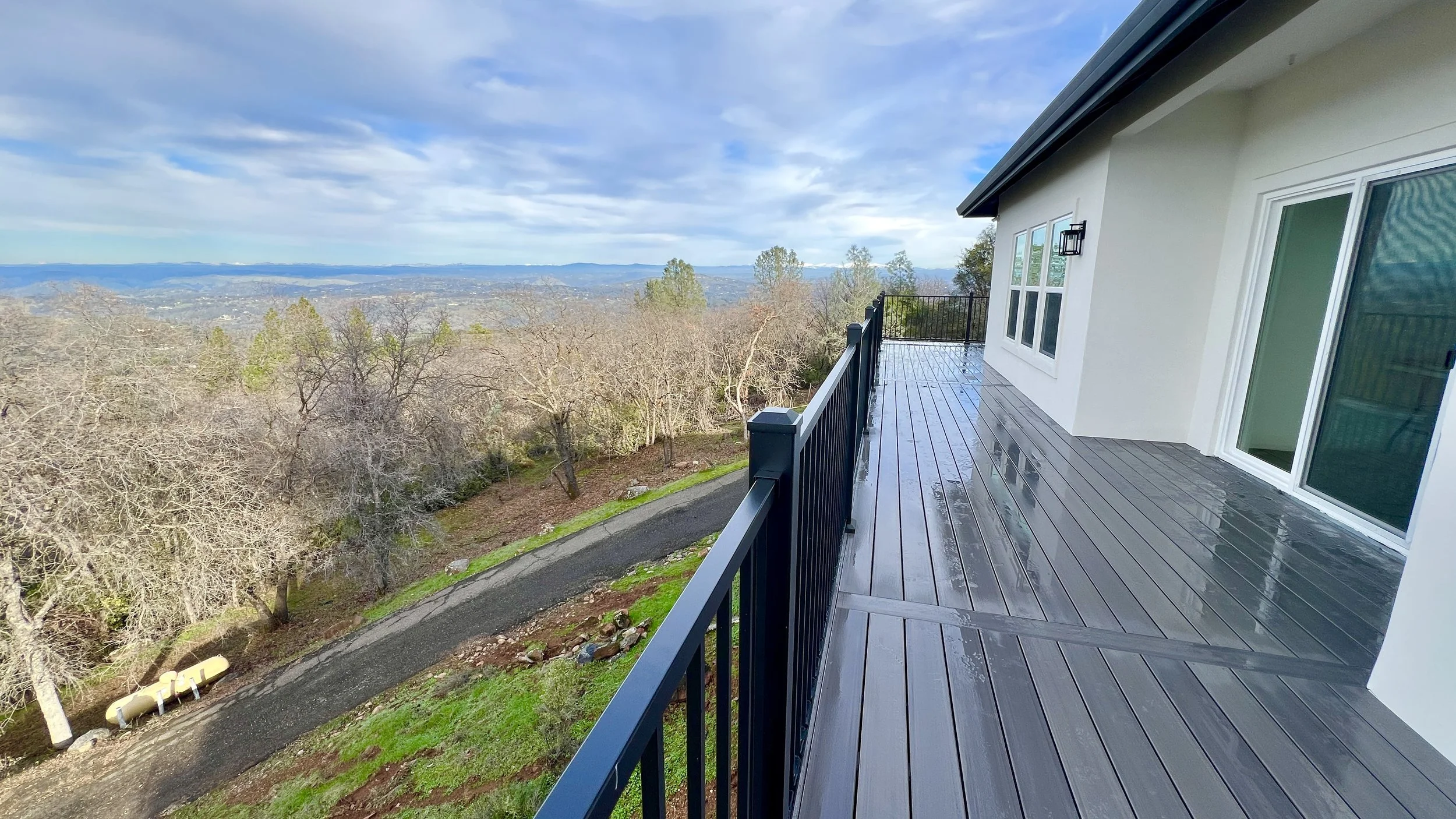 View from a house balcony showing a mountainside landscape with trees, a paved pathway, and distant mountains under a partly cloudy sky.