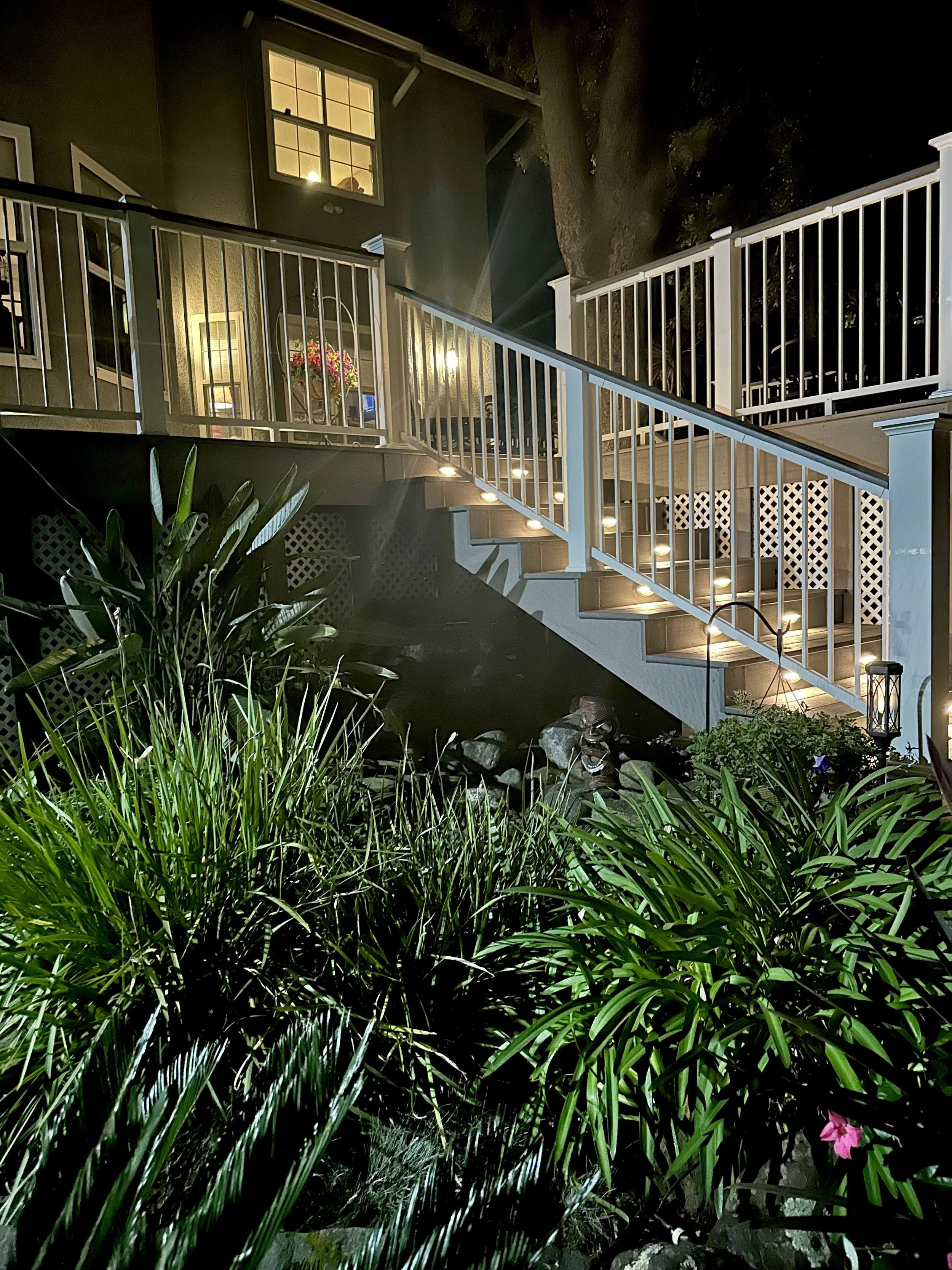 Nighttime view of an illuminated outdoor staircase with white railings, leading up to a house with lit windows. The staircase has small lights along the steps. In the foreground, there are lush green plants and a garden with rocks, a lantern, and add