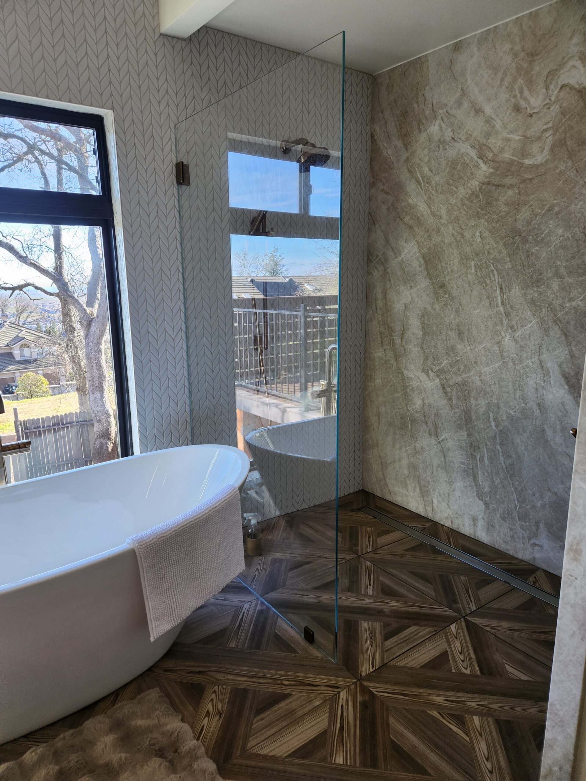 Bathroom with a freestanding bathtub, a large window, a glass shower partition, patterned wall tiles, and wood-patterned floor tiles.