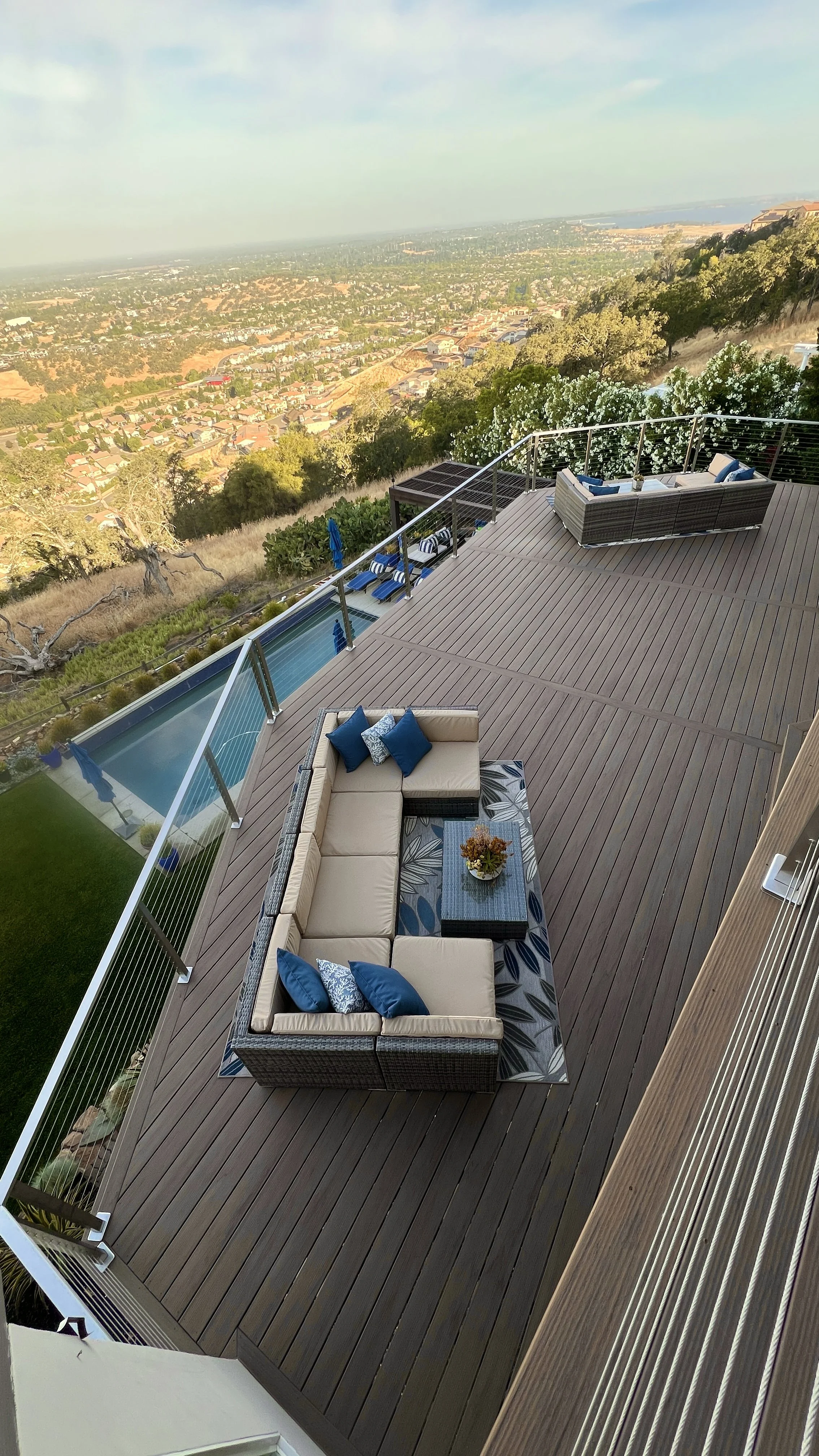 View of a wooden deck with outdoor sofa, coffee table, and pool overlooking a scenic landscape with houses and trees.